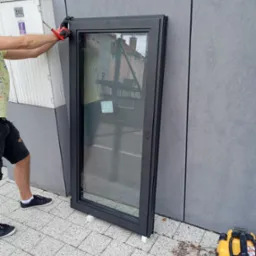 Czarna rama okienna oparta o szarą ścianę budynku, zabezpieczona białymi podkładkami, mężczyzna w czarnych rękawicach i koszulce w jasne wzory przykręca śrubokrętem górny zawias, w tle widoczny...