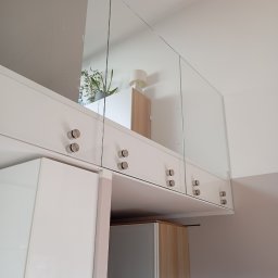 NI-TECH ROMAN Jedenasty - Nowoczesne szafki wiszące z białymi frontami i szklanymi drzwiczkami mocowanymi na ozdobnych, okrągłych uchwytach, z widoczną zawartością szafek w tle.