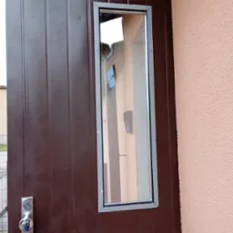 Brązowe drzwi z prostokątnym oknem, widok z bliska. W szkle odbija się fragment budynku. Metalowa klamka po lewej stronie. Część ściany po prawej.