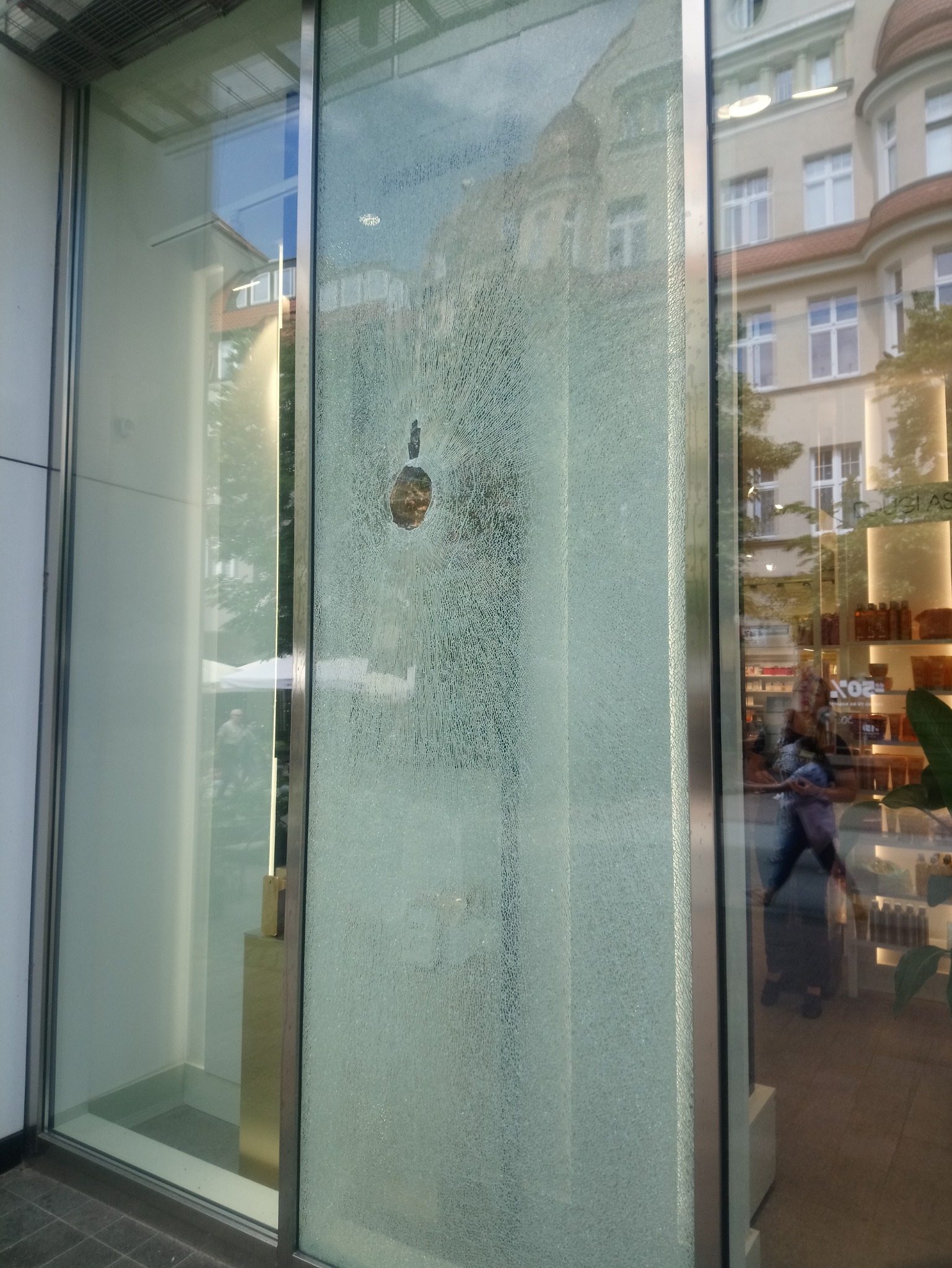 Rozbita szyba wystawowa z widocznym otworem po uderzeniu i promienistymi pęknięciami, odbicie ulicy i wnętrza sklepu.