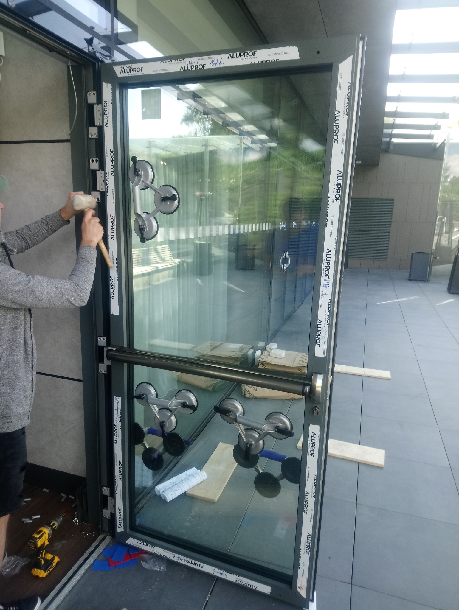 Montaż szklanej ściany działowej z użyciem przyssawek i młotka gumowego, widoczne narzędzia i materiały pomocnicze.