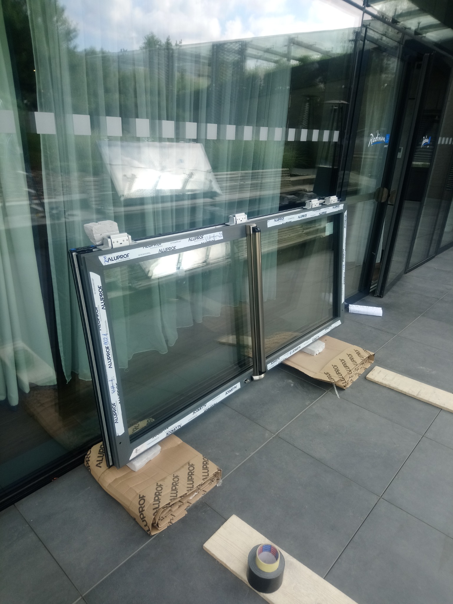 Dwa okna aluminiowe marki Aluprof, oparte o kartony i drewniane deski, czekają na montaż w budynku z widocznymi szklanymi fasadami i logo Radisson.