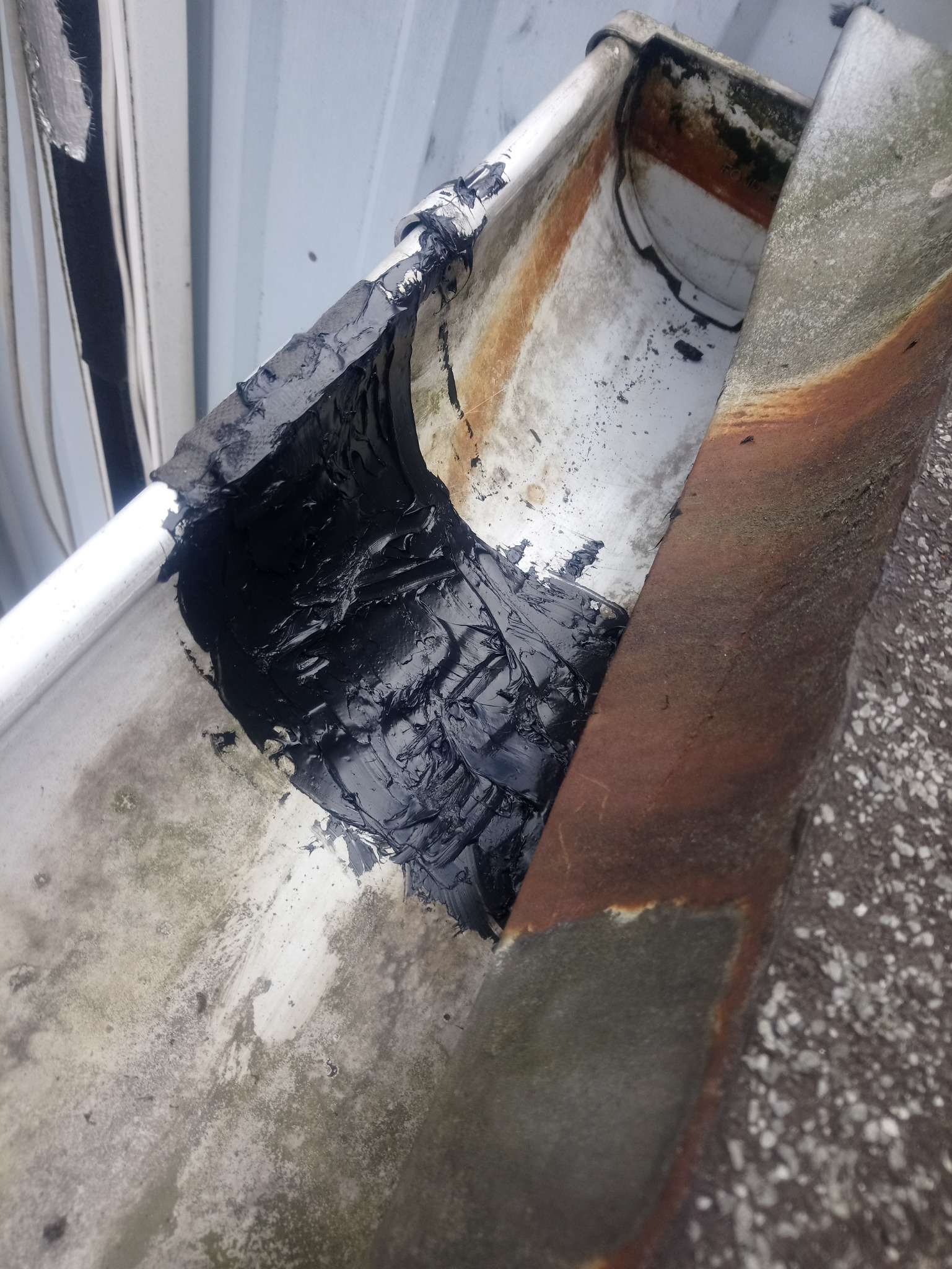 Szczegółowe ujęcie naprawianej rynny z użyciem czarnej masy uszczelniającej, widoczne ślady korozji i zabrudzeń na metalowej powierzchni.