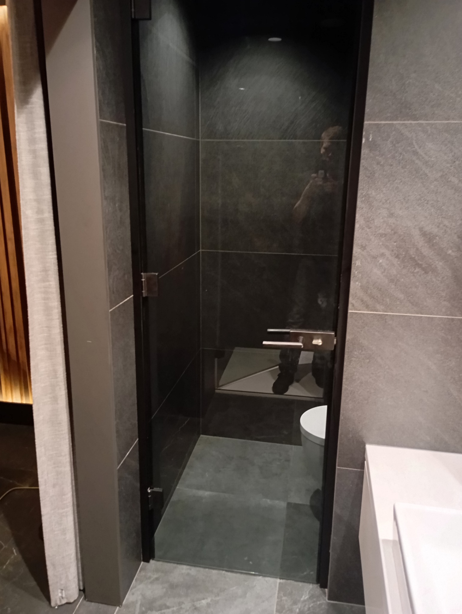 Szklana kabina prysznicowa z ciemnymi płytkami, widoczne zawiasy i uchwyt, odbicie osoby robiącej zdjęcie.