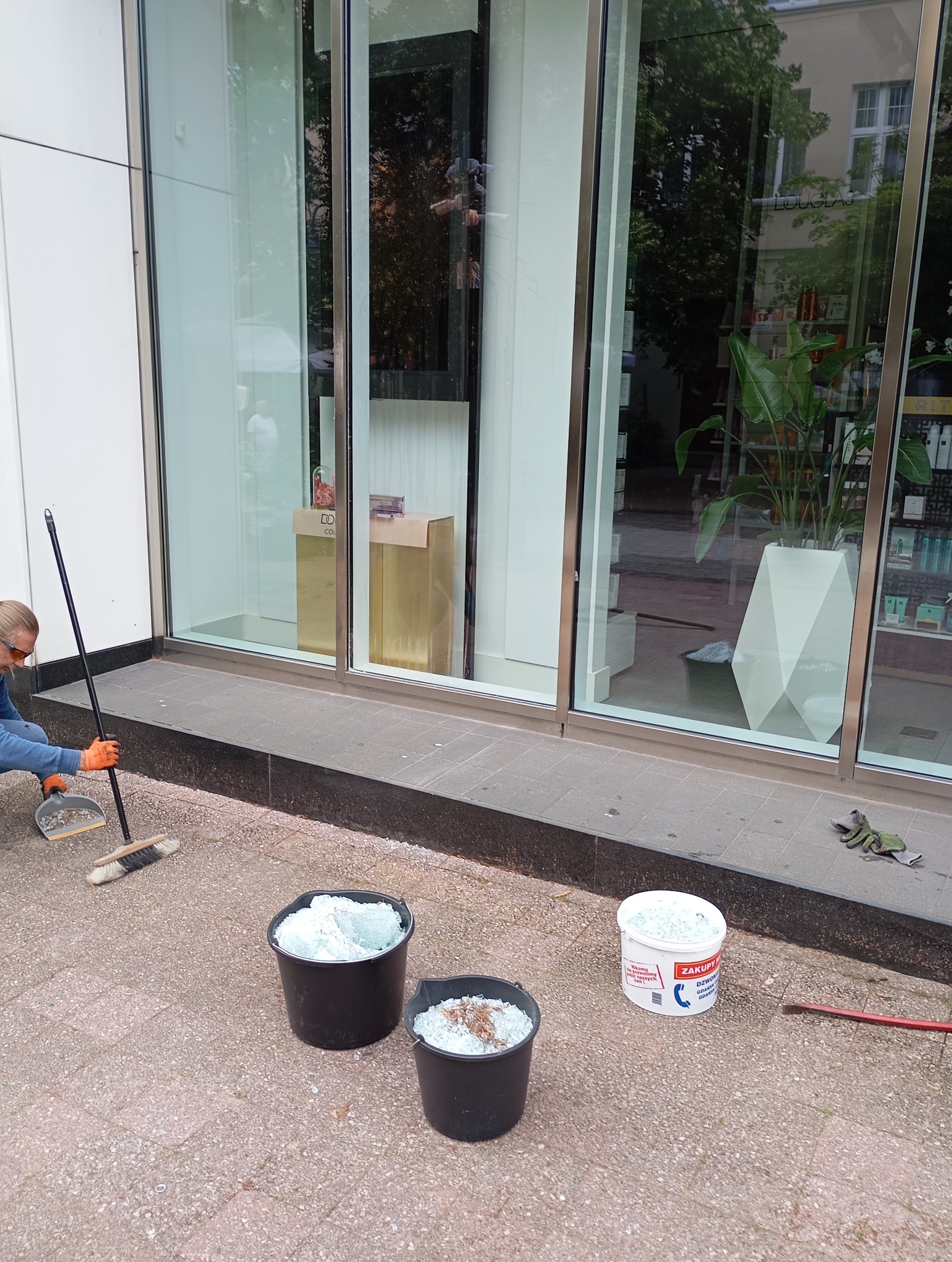 Mężczyzna w pomarańczowych rękawicach sprząta potłuczone szkło przed witryną sklepową, używając zmiotki i szufelki; obok stoją wiadra wypełnione szkłem i puste opakowanie po zaprawie.