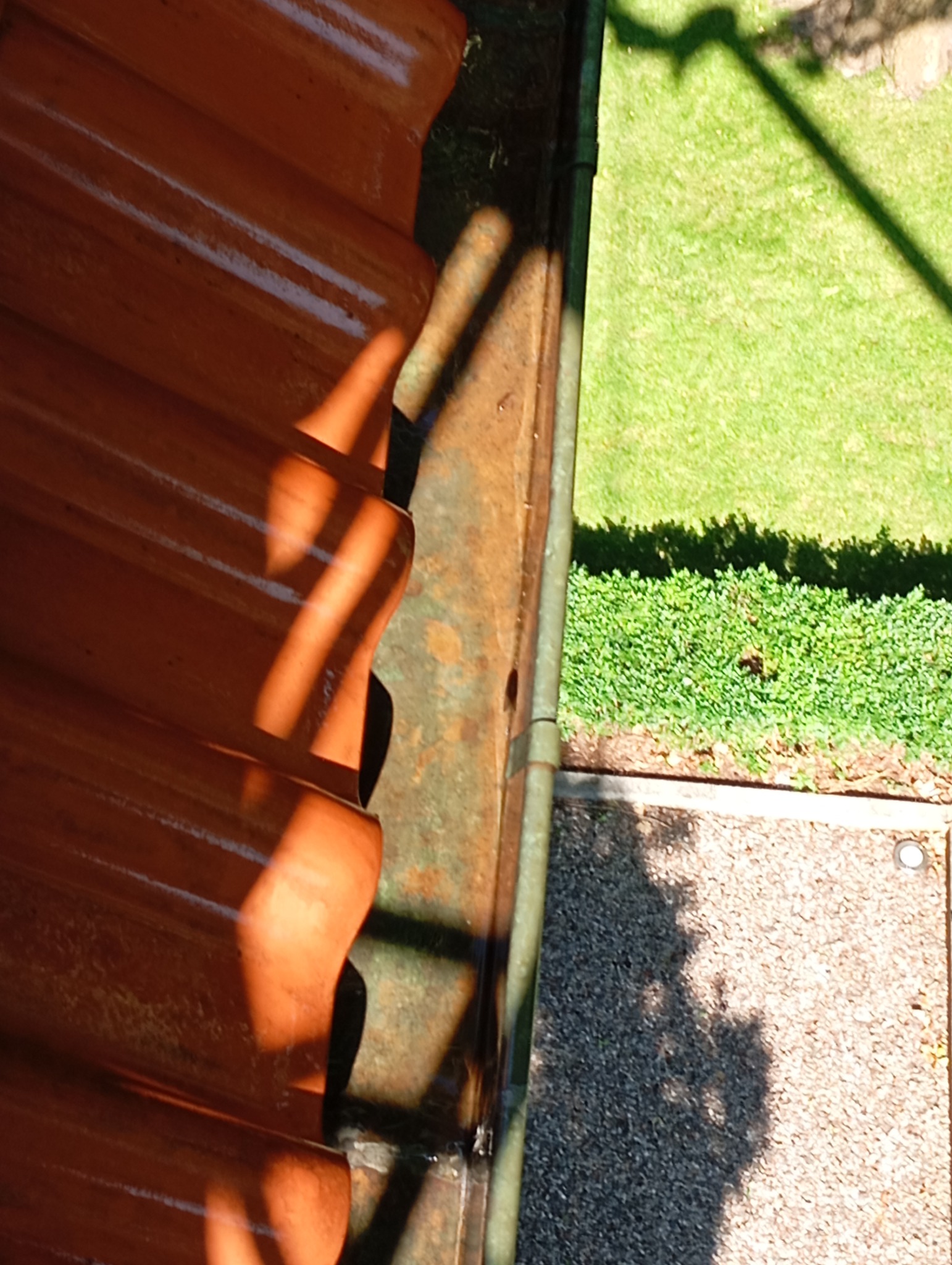 Fragment dachu z pomarańczową dachówką i metalową rynną, widoczne z góry, z fragmentem trawnika i ścieżki z kamyczków w tle.