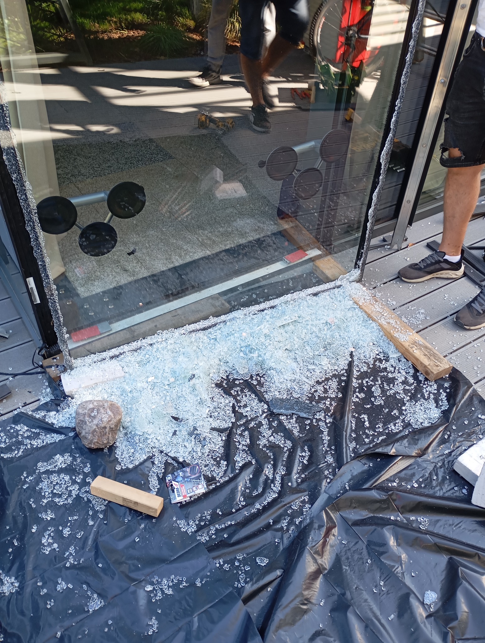 Rozbite szkło z okna, zabezpieczone czarną folią, z przyssawkami do szkła widocznymi na ramie okna, oraz fragmentem drabiny i narzędzi w tle.