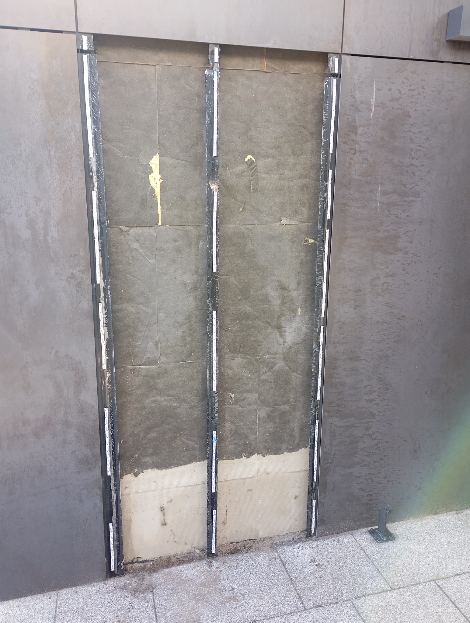 Przygotowanie otworu w ścianie pod montaż drzwi szklanych, widoczne listwy montażowe i fragment wypełnienia otworu, szara ściana zewnętrzna.