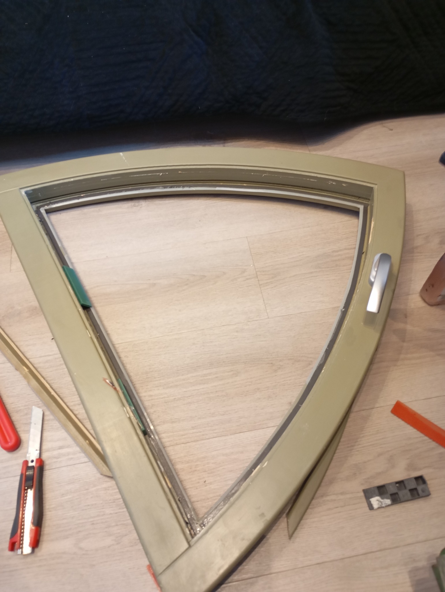 Nietypowe okno trójkątne w trakcie montażu, widoczne narzędzia: nożyk tapicerski oraz podkładki dystansowe, całość leży na podłodze.