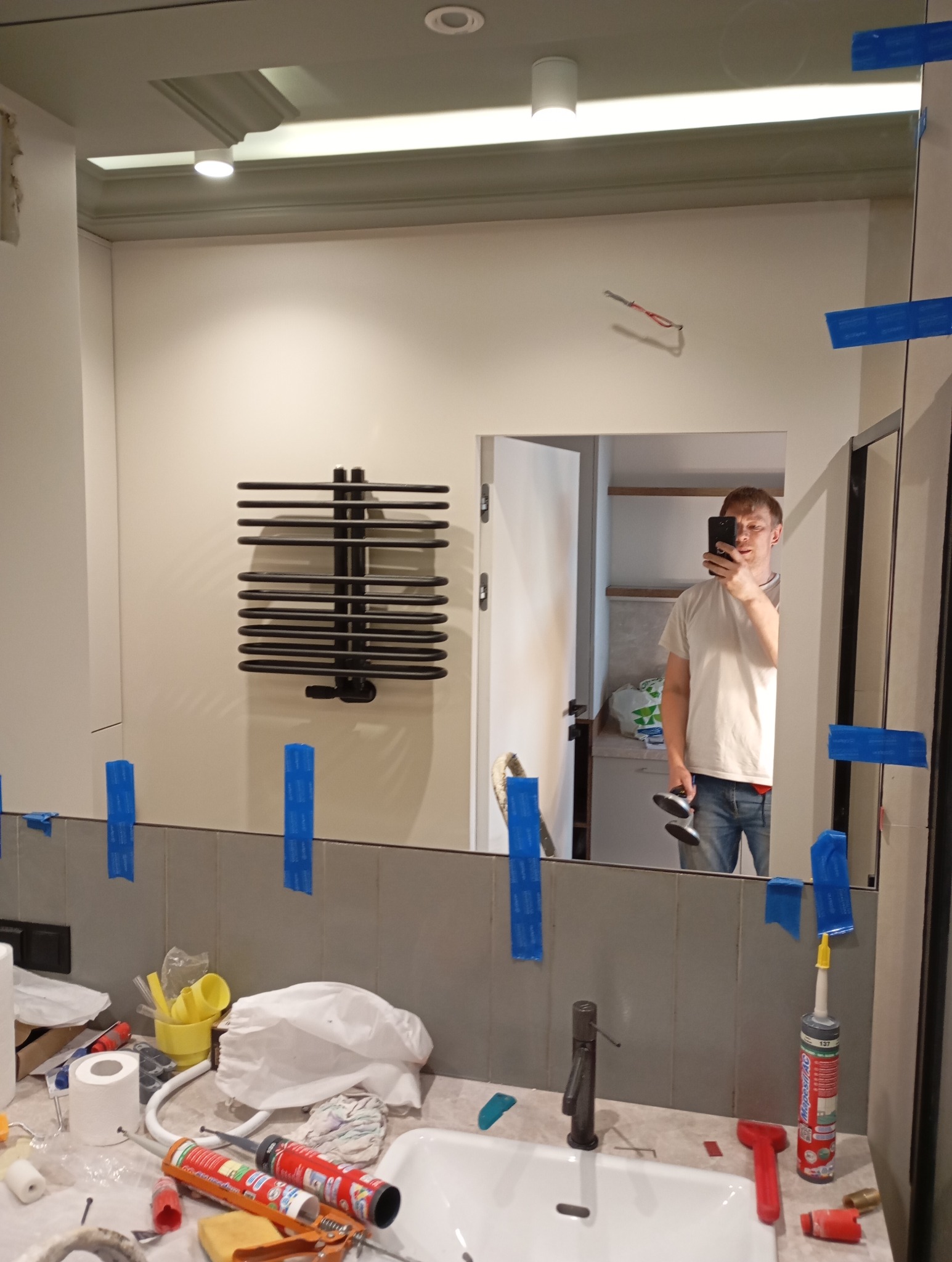 Montaż lustra w łazience, zabezpieczonego niebieską taśmą malarską. W tle widoczne narzędzia, umywalka i grzejnik drabinkowy. Odbicie osoby robiącej zdjęcie.