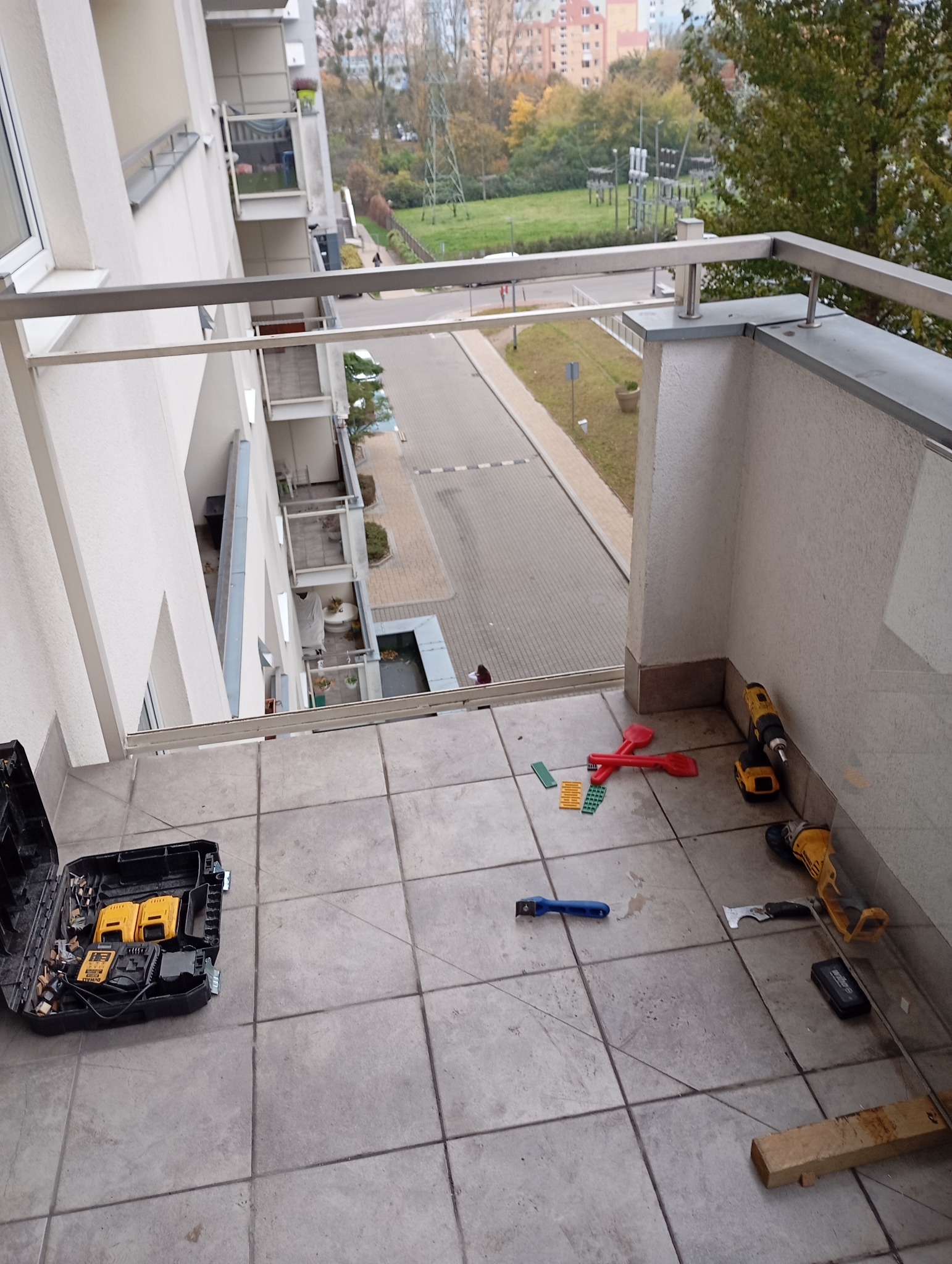 Balkon w bloku z narzędziami do montażu balustrady. Widoczne wiertarki, skrzynka narzędziowa i elementy mocujące. W tle widok na osiedle i zieleń.