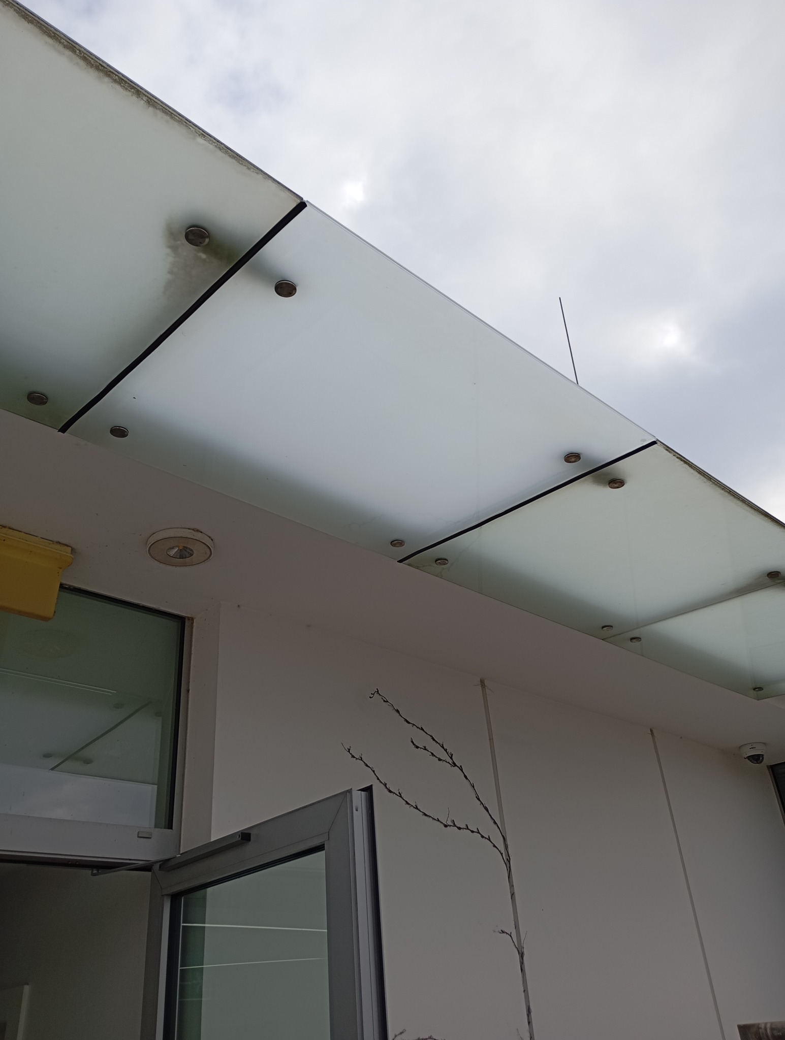 Szklany daszek nad wejściem do budynku, mocowany na metalowych śrubach. Widoczne drzwi wejściowe i fragment ściany z dekoracyjnym motywem gałązek. Pochmurne niebo w tle.