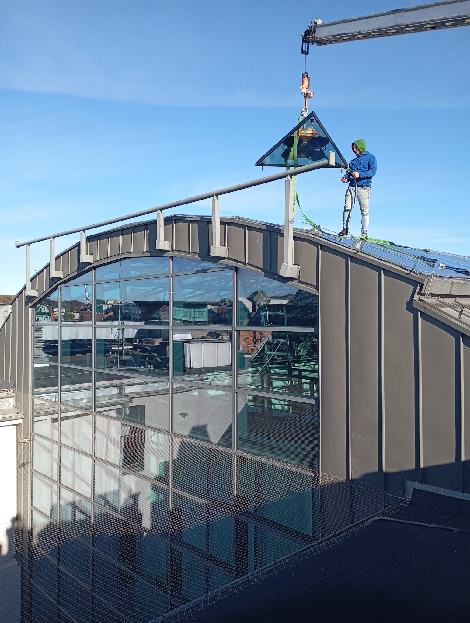 Montaż trójkątnej szyby na dachu budynku z użyciem dźwigu. Pracownik w niebieskiej kurtce zabezpiecza szybę. Widoczne duże okna budynku i błękitne niebo.