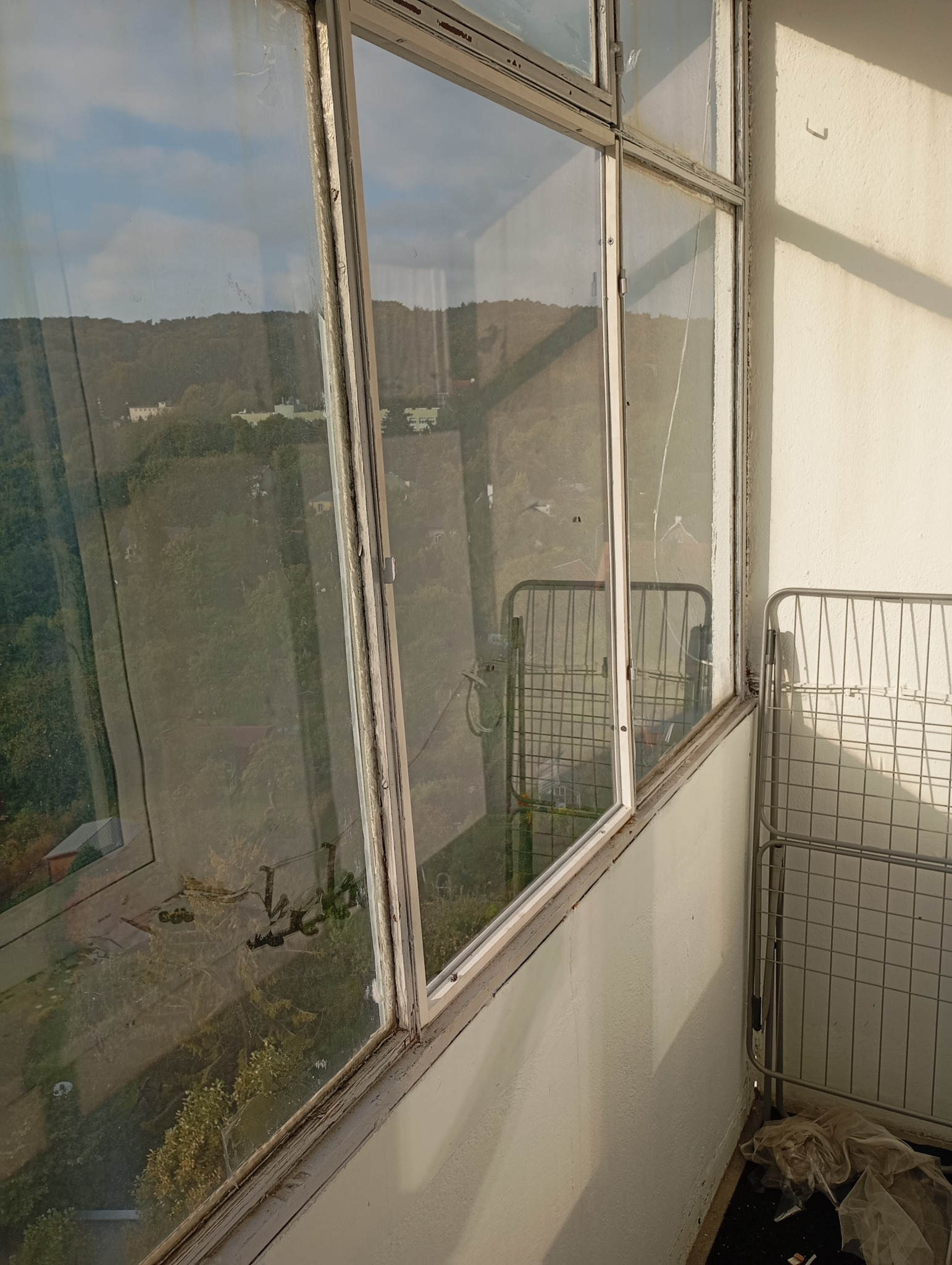Stare, brudne okna balkonowe z widokiem na zieleń. Widoczne ślady użytkowania i zaniedbania. W tle suszarka na pranie. Nierównomierne oświetlenie.
