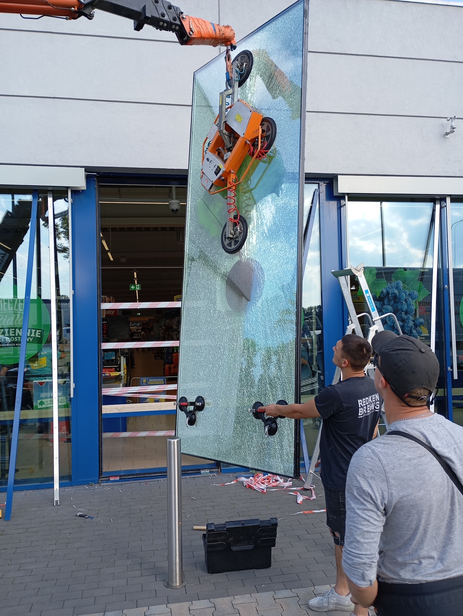 Montaż dużej szyby witrynowej w sklepie za pomocą przyssawki i dźwigu. Widoczni dwaj monterzy, narzędzia i taśma ostrzegawcza. Praca szklarza w trakcie realizacji.