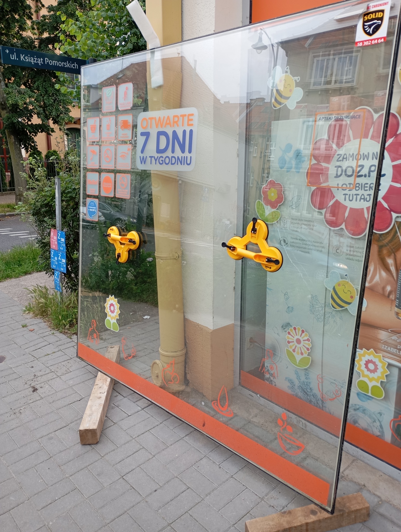 Szyba okienna z przyssawkami na chodniku, oparta o drewniane klocki. Na szybie naklejki i reklamy apteki. Widoczny znak uliczny.