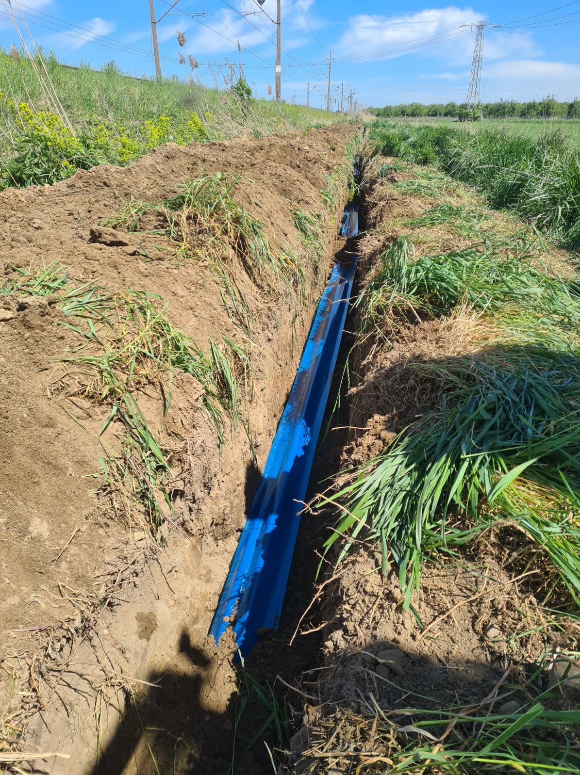 Wykop w ziemi z ułożonym niebieskim rurociągiem, widoczne trawy i słupy energetyczne w tle.