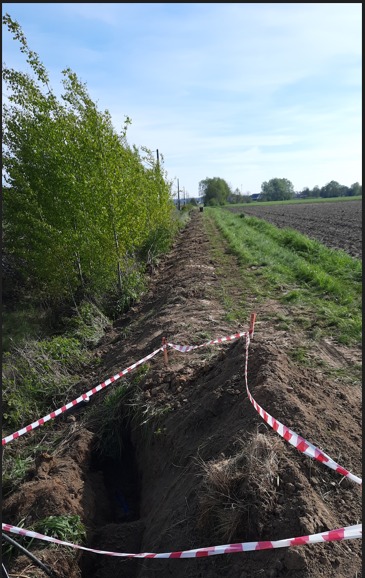 Długi wykop wzdłuż drzew, zabezpieczony taśmą ostrzegawczą, z widocznymi kablami wewnątrz, na tle zaoranego pola i błękitnego nieba.