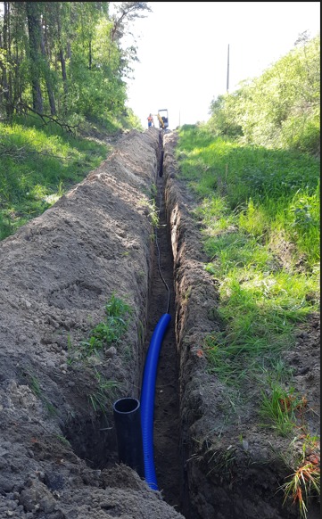 Długi, wąski wykop w ziemi z ułożoną niebieską rurą karbowaną i czarną rurą PCV, w tle widoczny operator koparki i las.