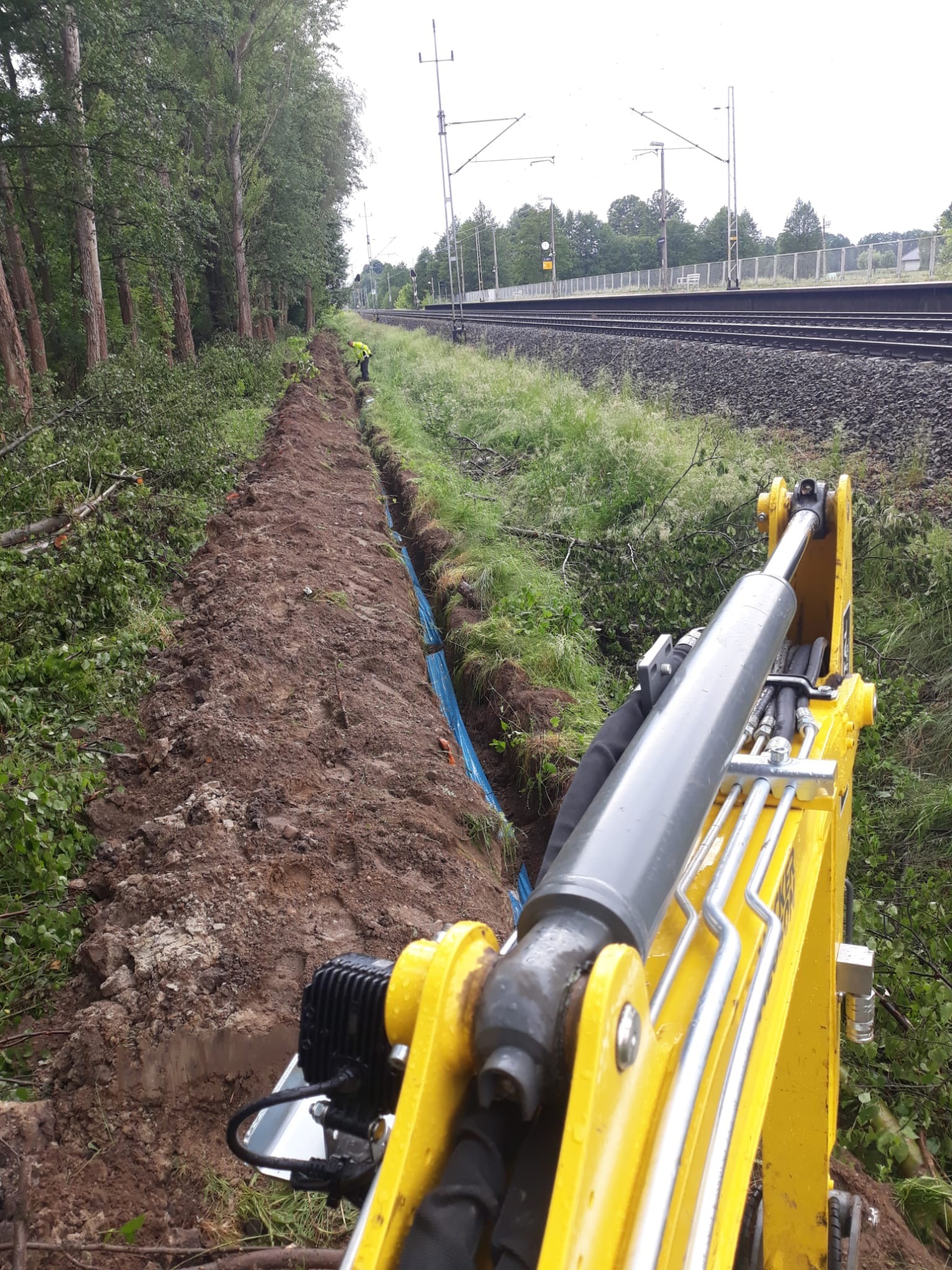Wykopany wąski rów z niebieską rurą na dnie, widoczny z perspektywy kabiny żółtej koparki, w tle linia kolejowa i pracownik w odblaskowej kamizelce.