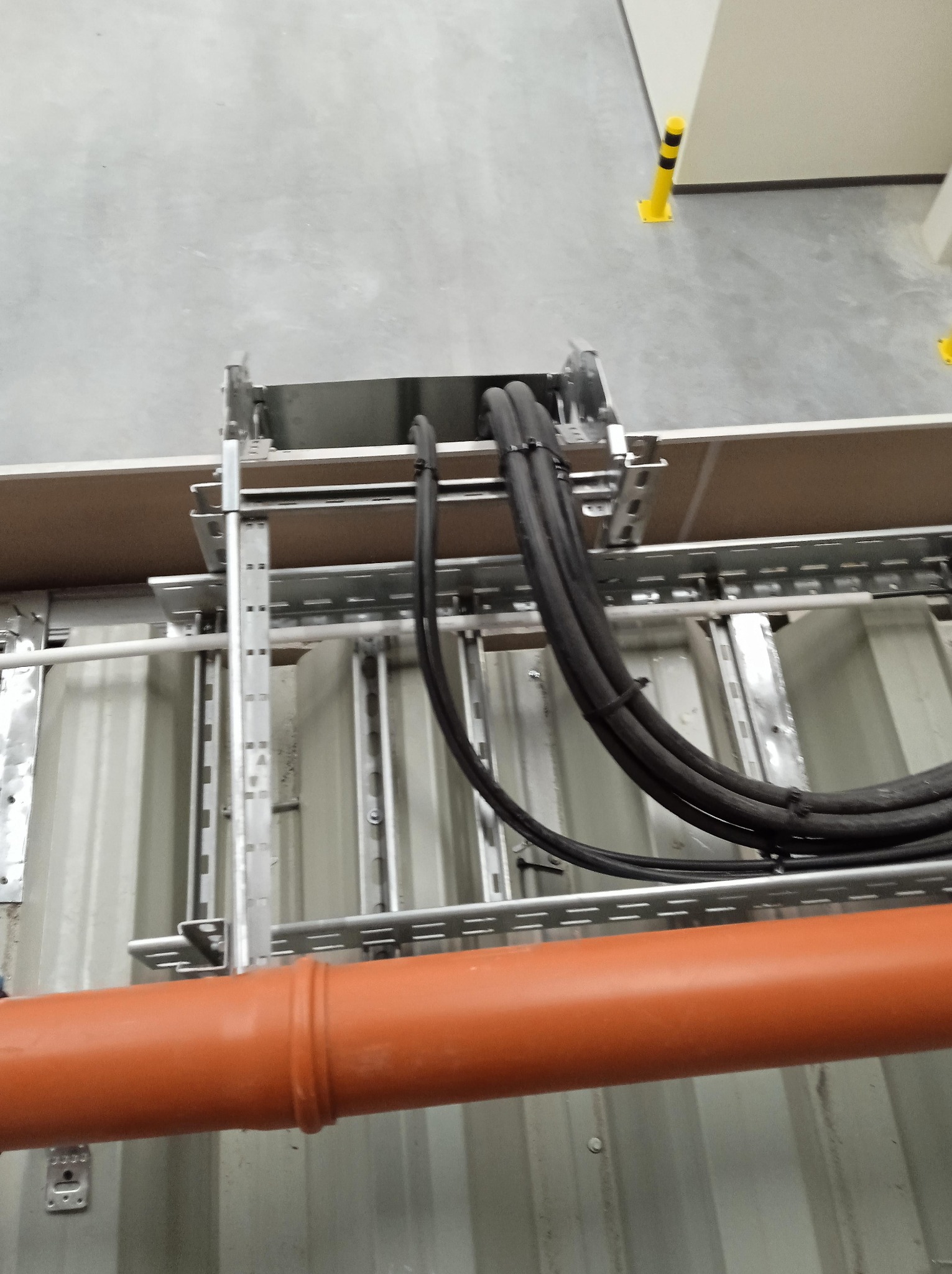 Metalowa drabinka kablowa z zamocowanymi czarnymi kablami i pomarańczową rurą, widoczne zabezpieczenia w tle.