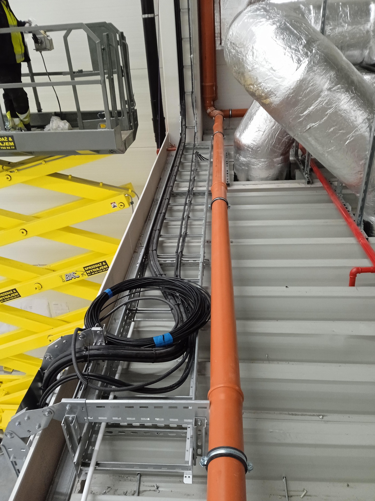 Montaż instalacji telekomunikacyjnych w hali przemysłowej: widoczne kable w korytach, rury wentylacyjne z izolacją termiczną, pomarańczowa rura instalacyjna oraz podnośnik nożycowy z pracownikiem...
