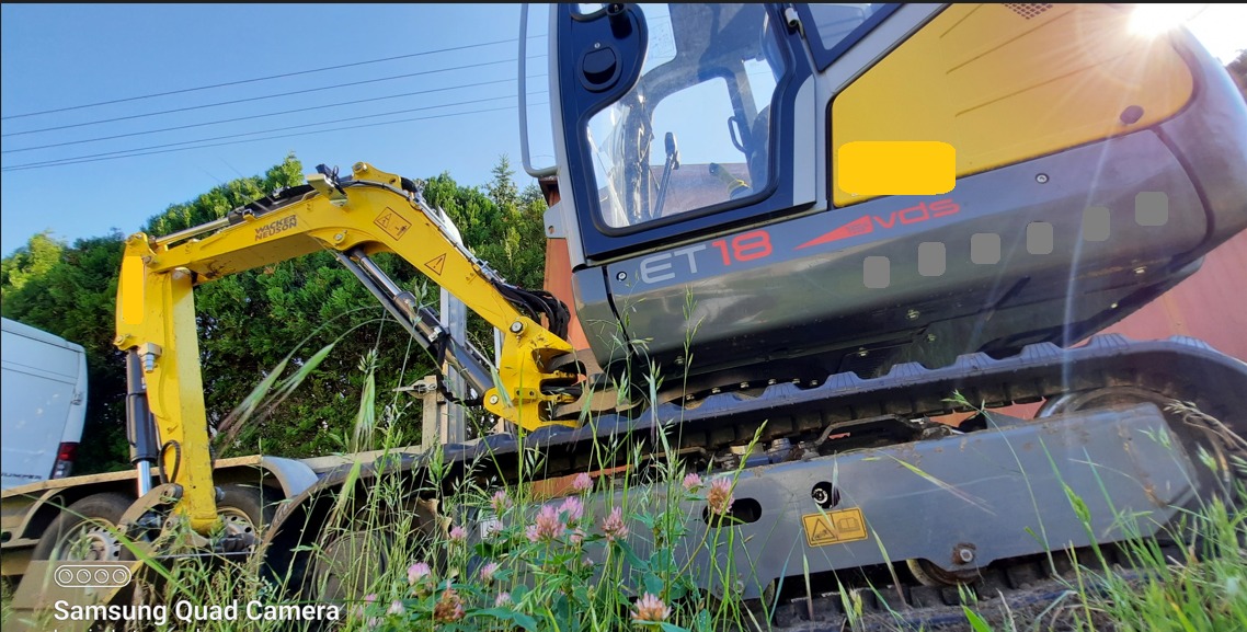 Żółto-szara minikoparka Wacker Neuson ET18 na przyczepie, widoczna z boku, z wysuniętym ramieniem, na tle zieleni i błękitnego nieba.