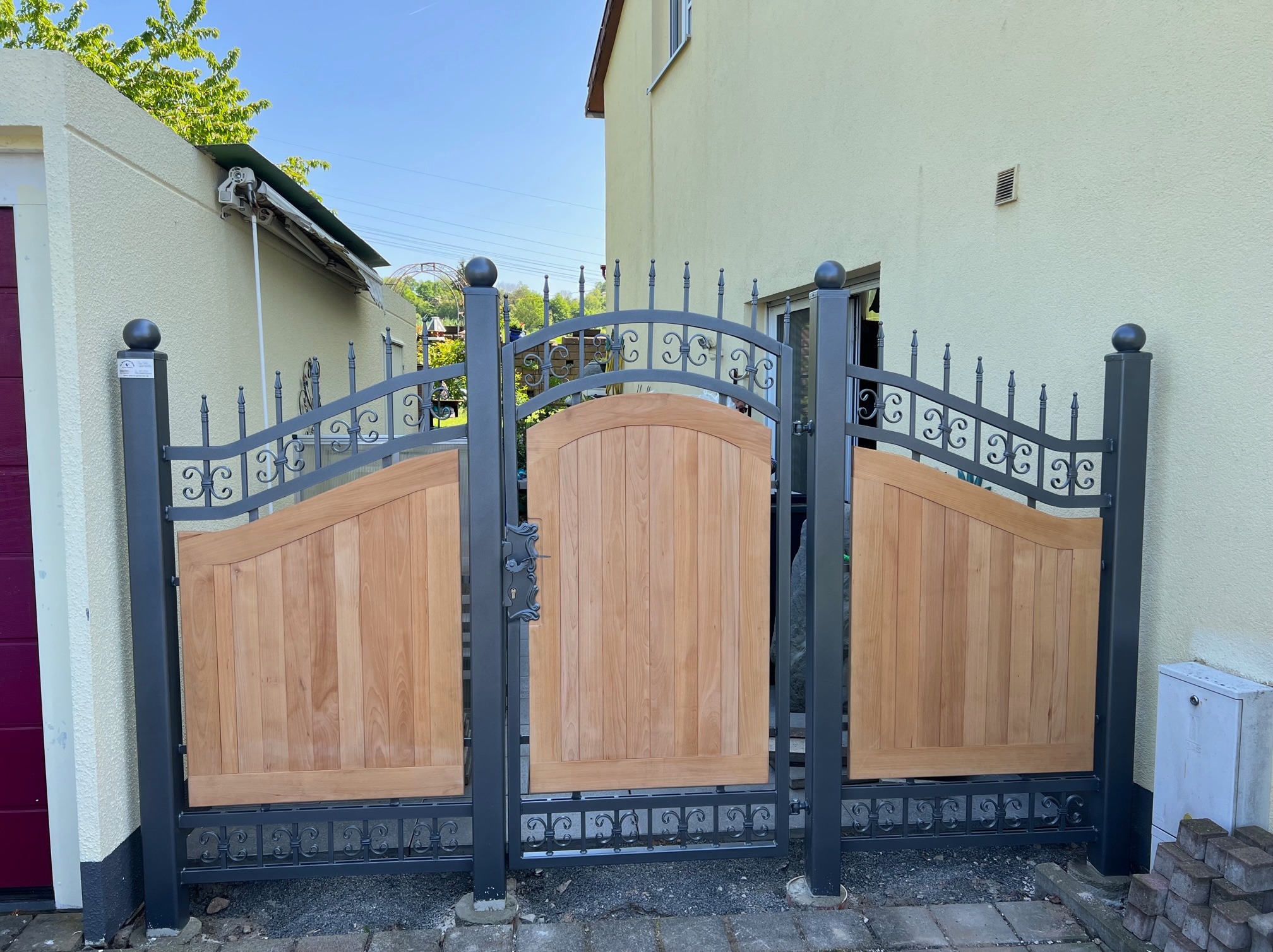 Nowoczesna brama wjazdowa z elementami drewnianymi i metalowymi, ozdobne detale na tle zabudowań mieszkalnych.