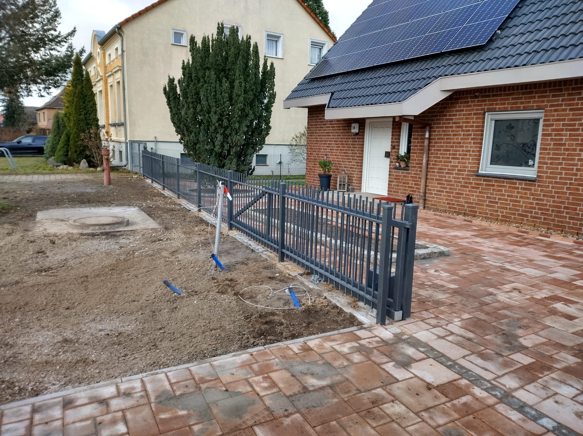 Szara metalowa furtka i ogrodzenie panelowe w trakcie montażu, widoczne narzędzia i przewody na ziemi, kostka brukowa i dom w tle.