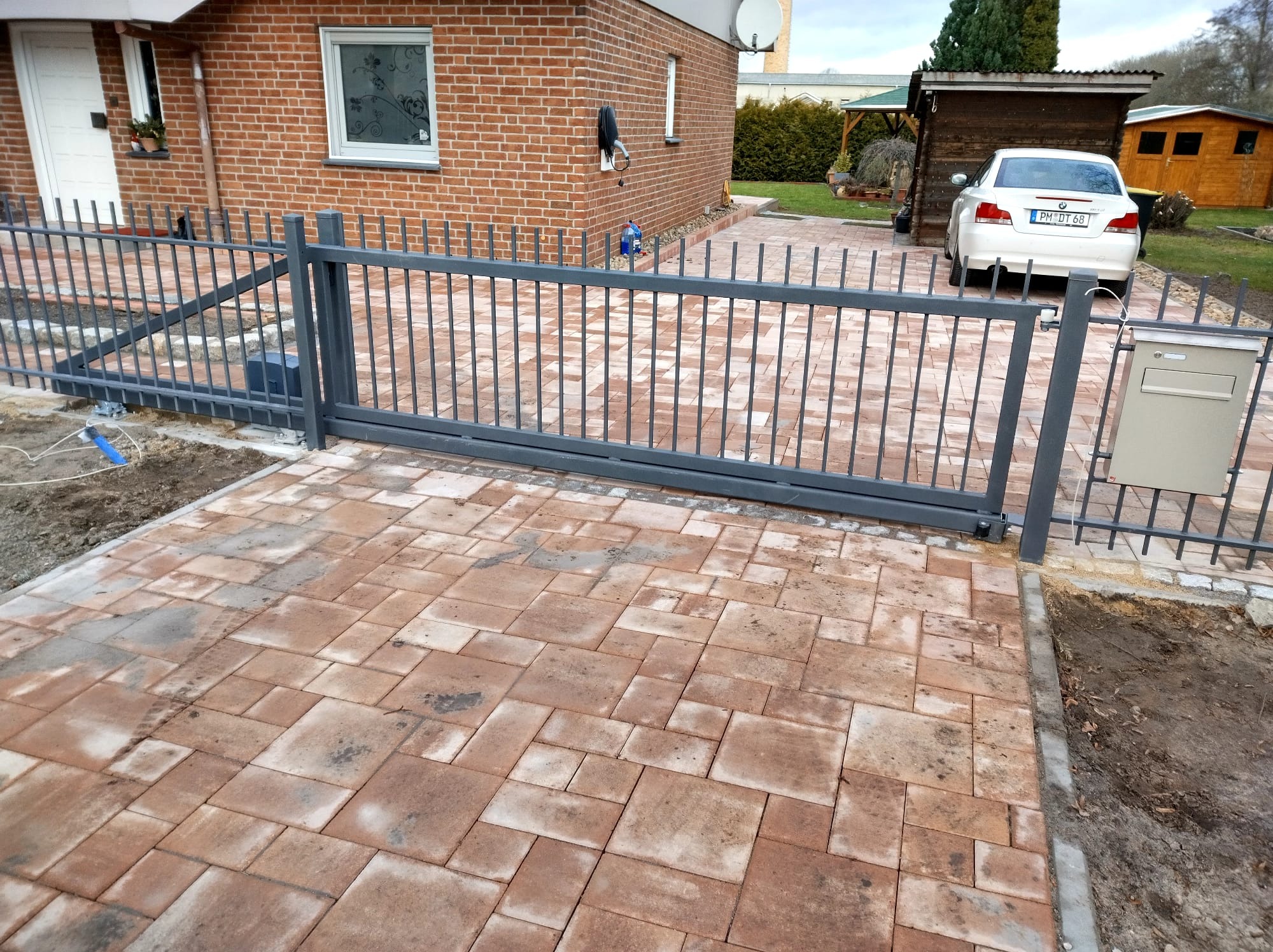 Nowoczesna, przesuwna brama wjazdowa z metalu, pomalowana na szaro, na posesji z wybrukowanym podjazdem z kostki w odcieniach czerwieni i brązu, widoczny dom z cegły w tle.