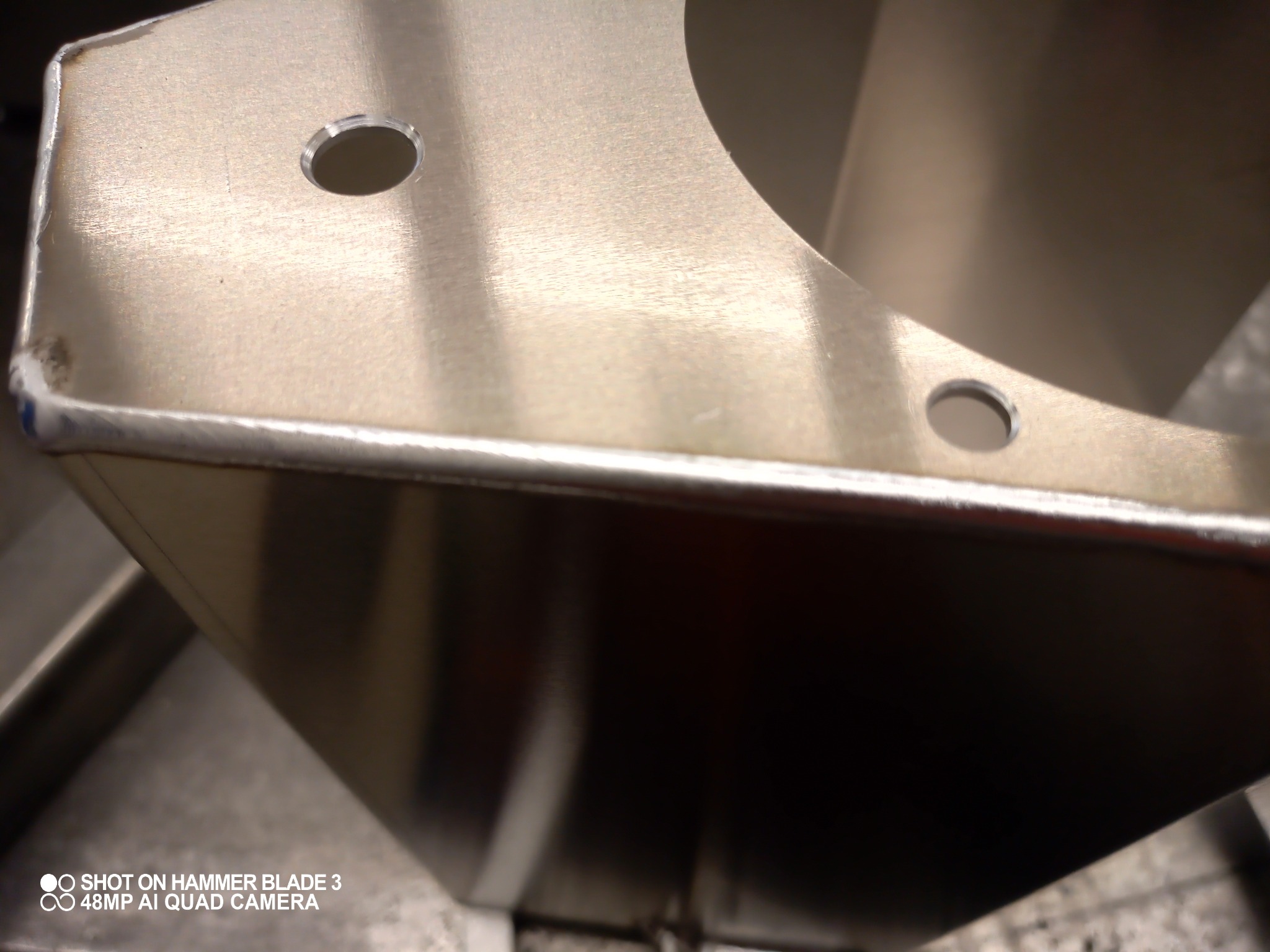 Metalowy element z widocznymi spawami na krawędziach i dwoma otworami, sfotografowany z bliska pod kątem.