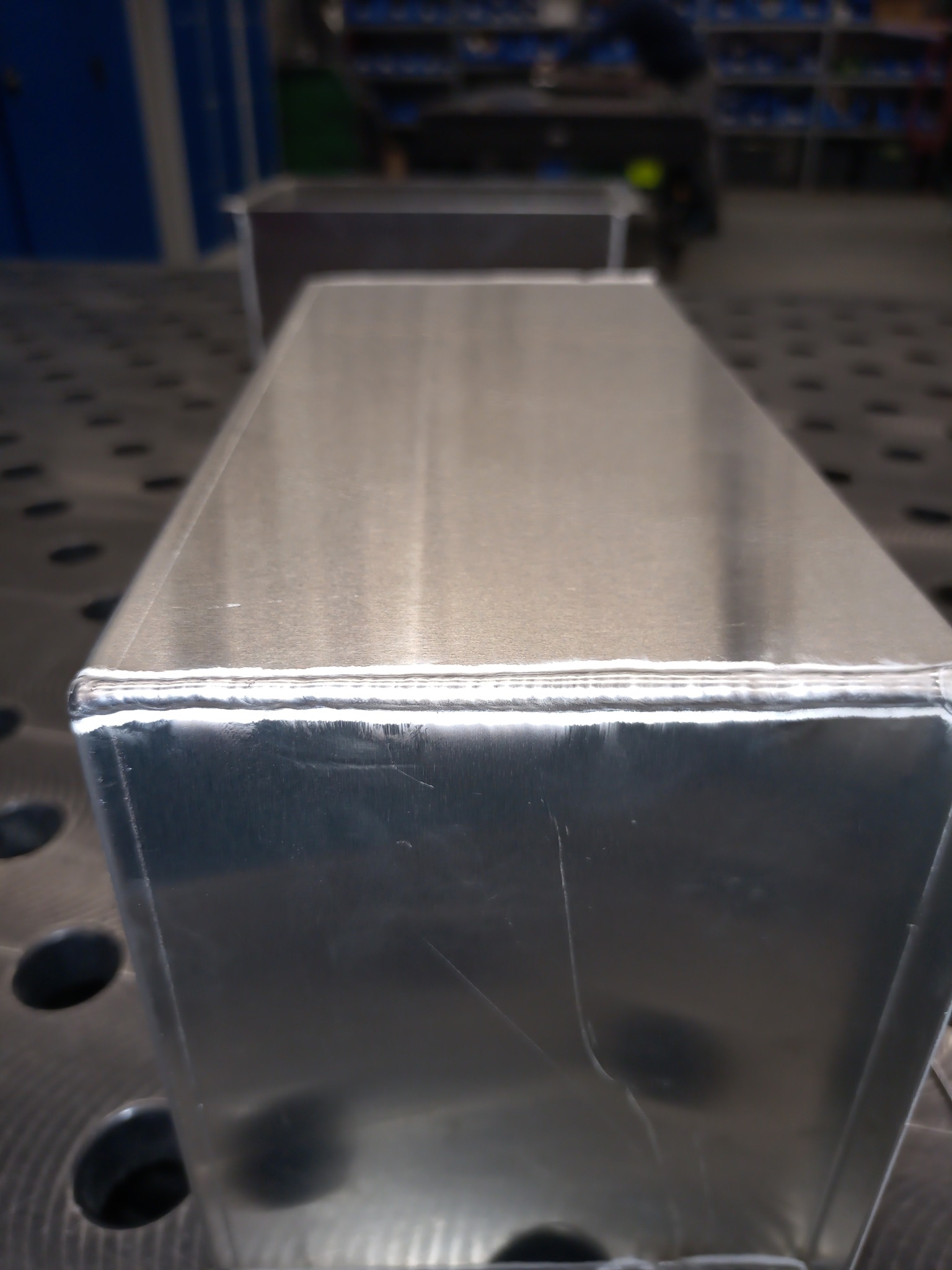 Prostopadłościenny pojemnik ze stali nierdzewnej z widocznymi spawami na krawędziach, umieszczony na perforowanym stole spawalniczym, w tle drugi, lekko rozmazany pojemnik.