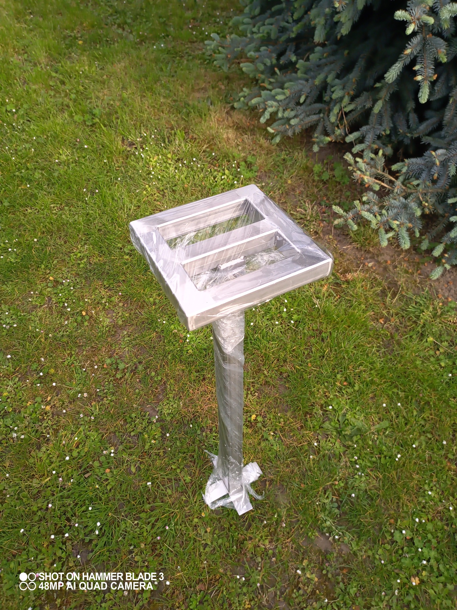 Metalowa konstrukcja spawana, owinięta folią ochronną, stojąca na trawie obok świerku.