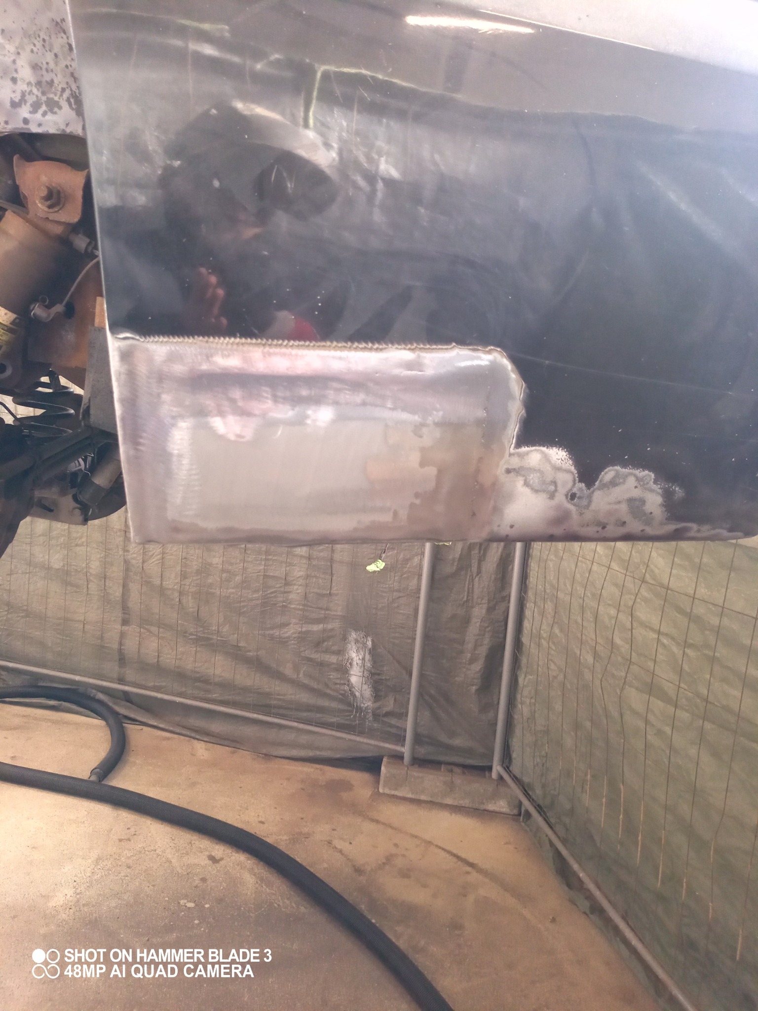 Fragment karoserii samochodu po wstawieniu nowej, metalowej blachy i wstępnej obróbce spoin, widoczne ślady szlifowania i przygotowania do dalszych prac blacharskich, tło stanowi zielona plandeka.