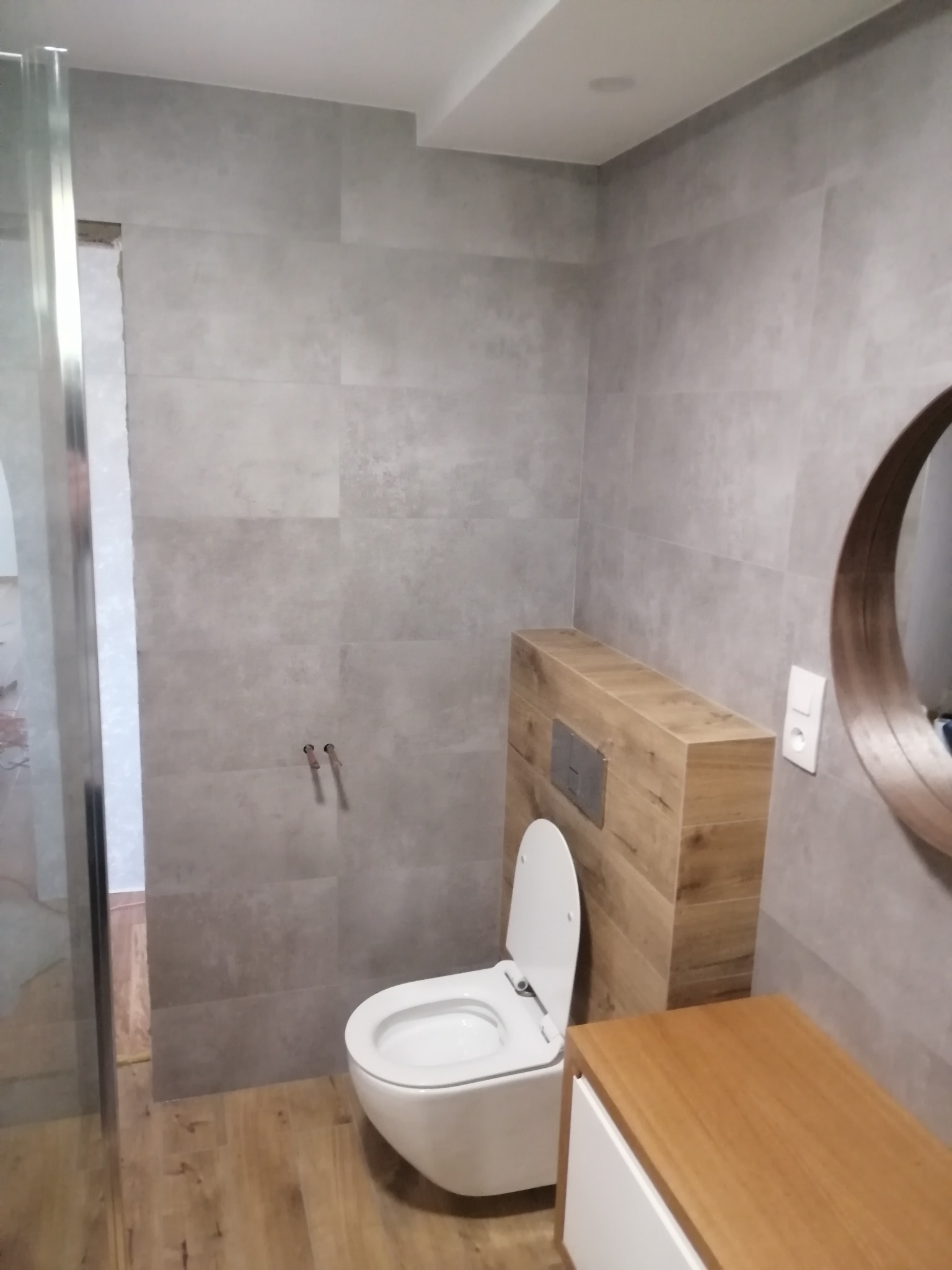 Nowoczesna łazienka z szarymi płytkami imitującymi beton, drewnopodobną podłogą i wiszącą toaletą z zabudową z drewnianych paneli, okrągłe lustro i drewniana szafka pod umywalkę.