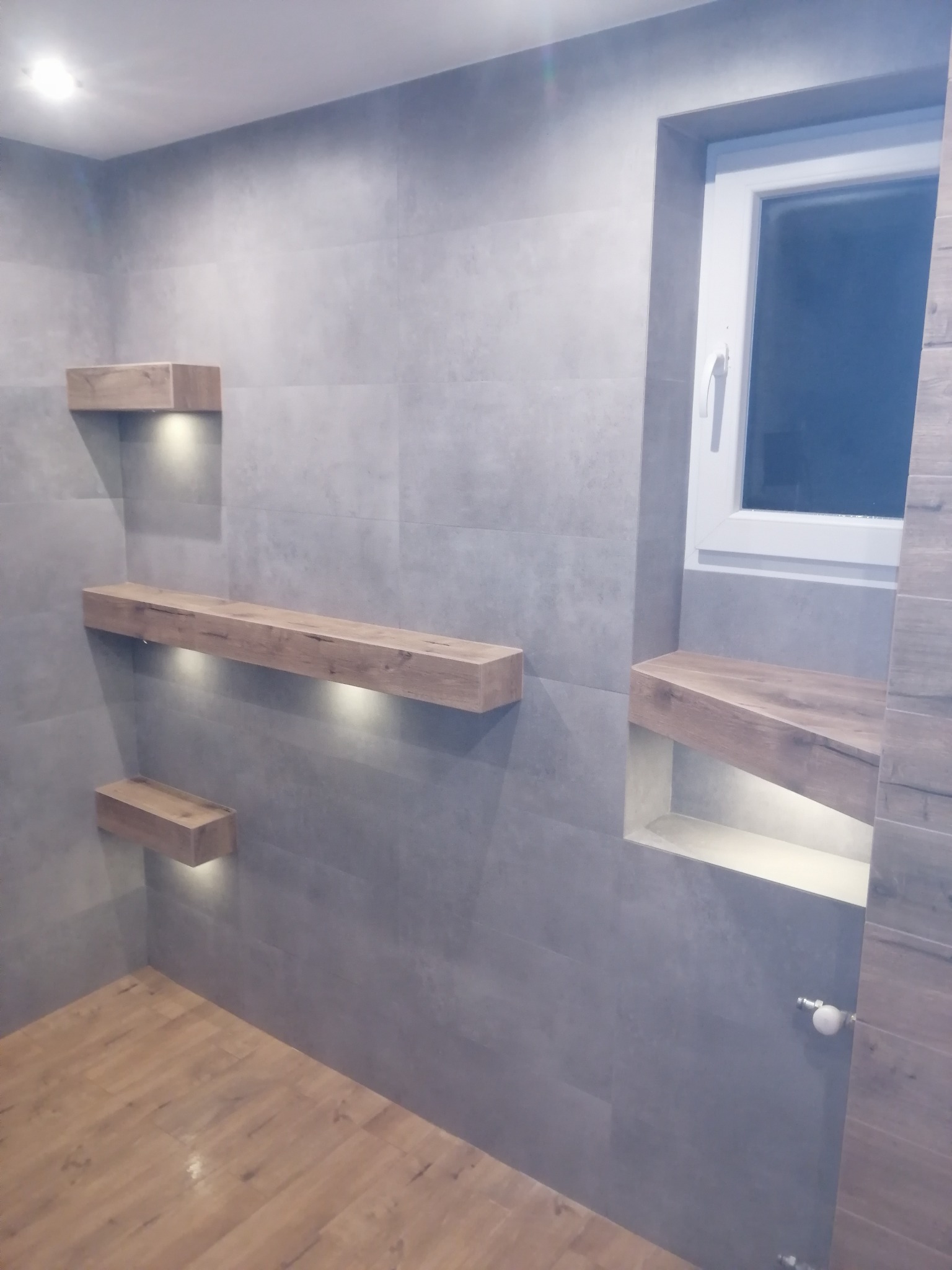 Wykończona łazienka z szarymi płytkami, drewnianymi półkami podświetlonymi punktowym światłem LED i oknem z białymi ramami.