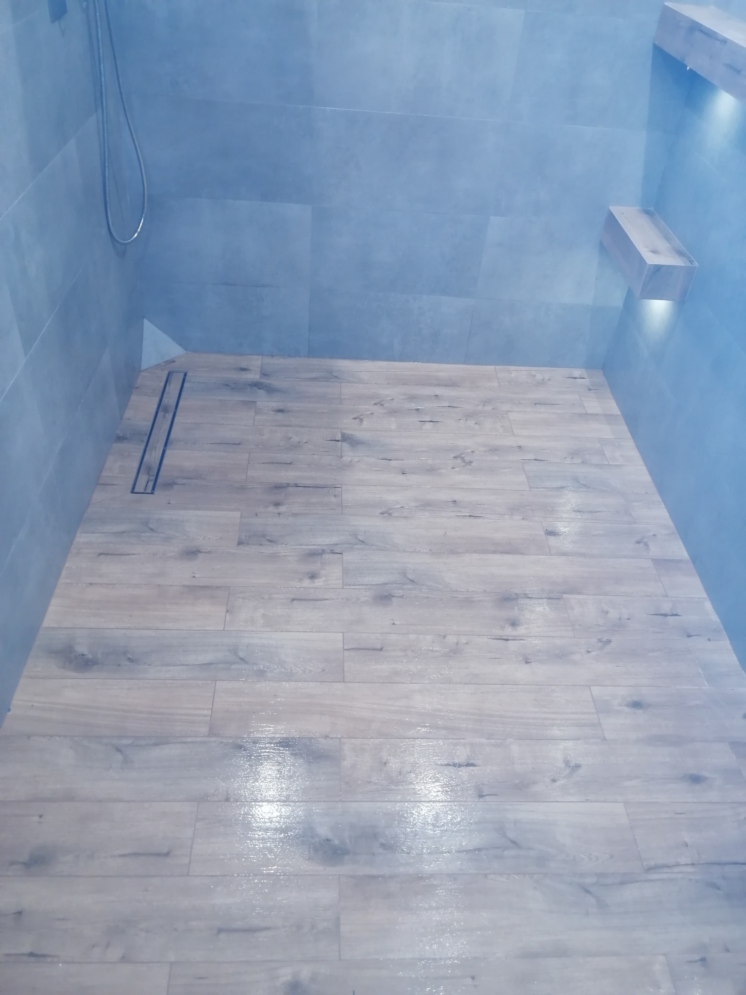 Nowoczesny prysznic walk-in z drewnopodobną podłogą, odpływem liniowym i szarymi płytkami ściennymi oraz półką z podświetleniem LED.