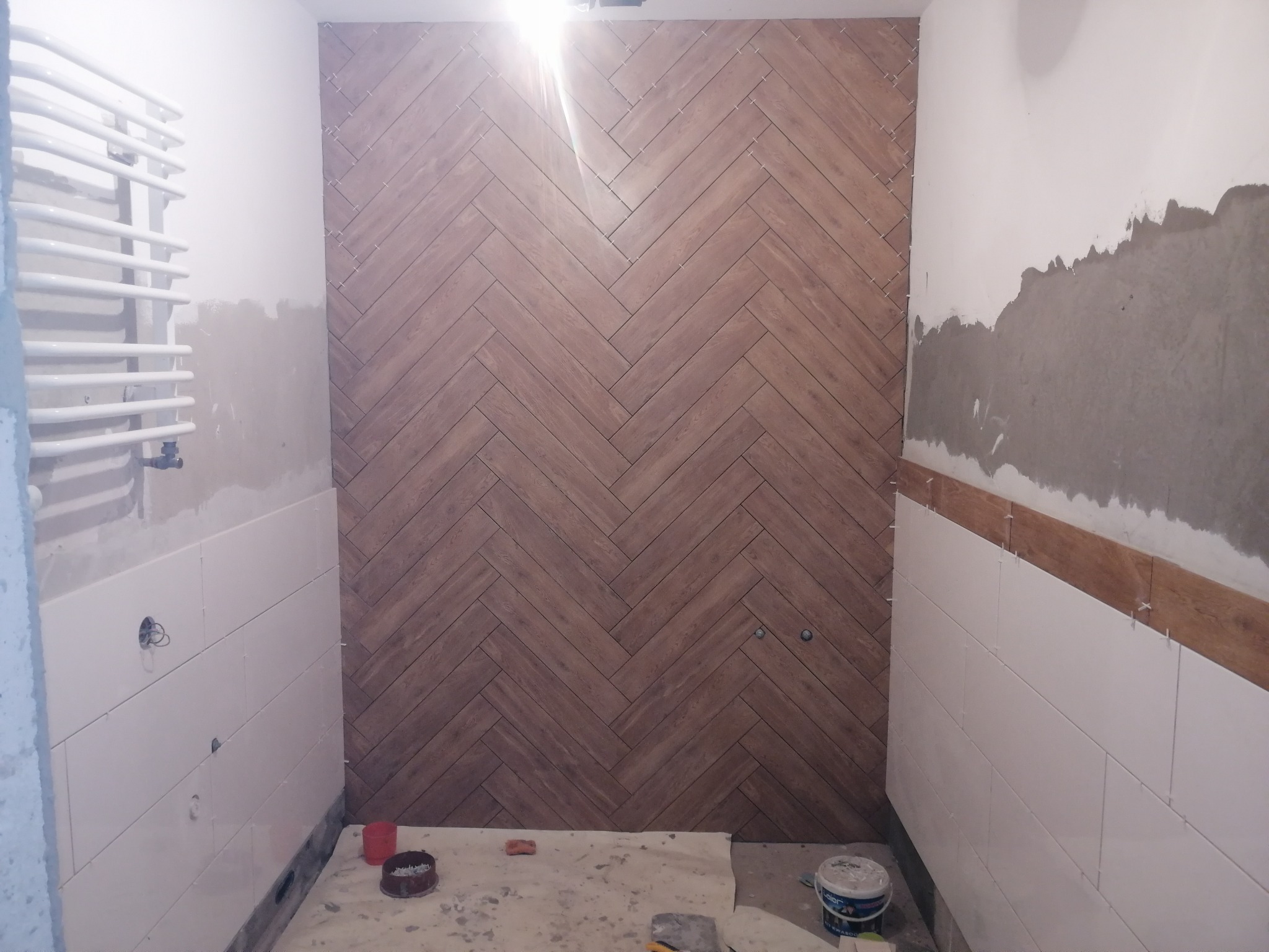 Łazienka w trakcie układania płytek: na jednej ścianie ułożone płytki drewnopodobne w jodełkę, na pozostałych ścianach białe płytki w trakcie montażu, widoczne krzyżyki dystansowe i pozostałości...