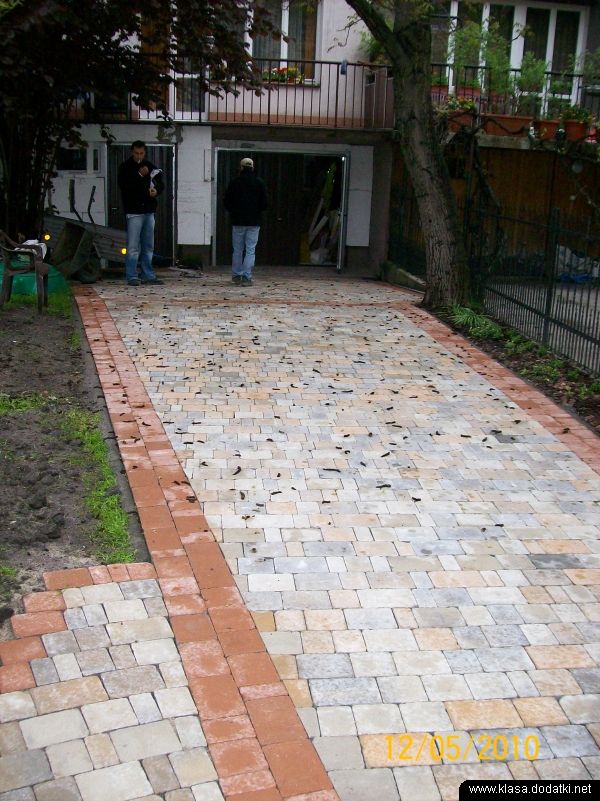 Ułożona kostka brukowa w odcieniach szarości i beżu z czerwoną obwódką prowadząca do garażu, dwóch mężczyzn w tle, liście na kostce