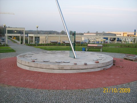 Zegar słoneczny z czerwoną kostką brukową wokół, widoczne niedokończone fragmenty układania kostki i elementy konstrukcyjne w tle.