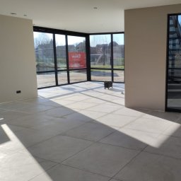 Skyhouse - Jasne wnętrze z ułożoną, kwadratową podłogą z płytek w odcieniach szarości, duże okna z czarnymi ramami wpuszczają dużo światła, widoczna reklama na zewnątrz, w rogu stoi mały wózek narzędziowy.