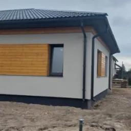 Nowoczesny dom jednorodzinny z szarą elewacją, drewnianymi panelami dekoracyjnymi pod oknami i ciemną dachówką, widoczny z zewnątrz na etapie wykończenia terenu.