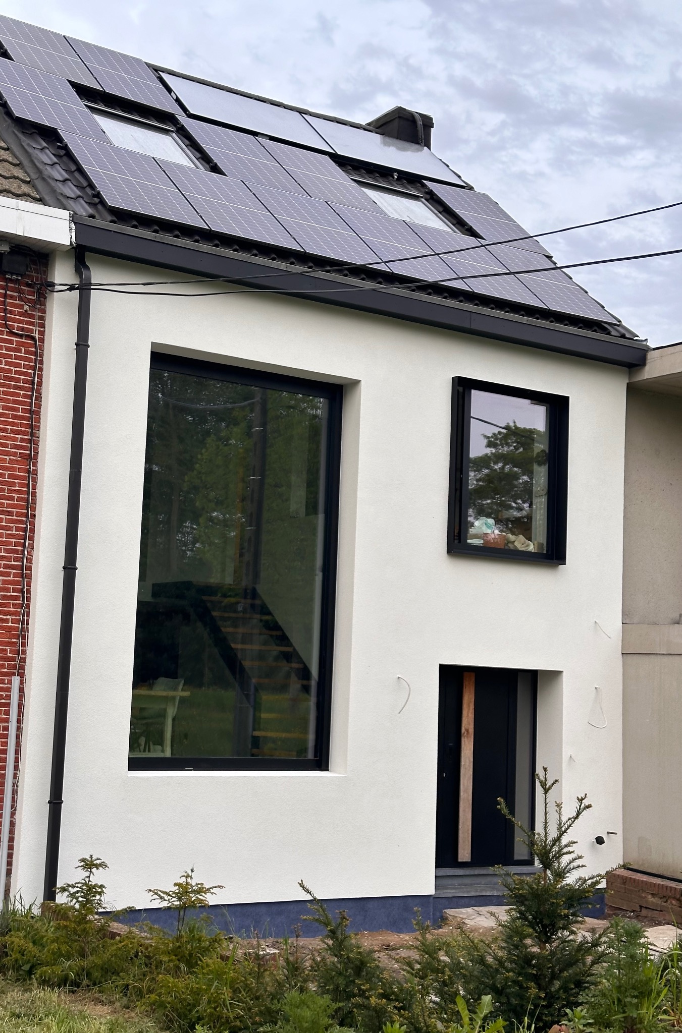 Biała elewacja domu z dużym oknem, mniejszym oknem i czarnymi ramami, czarne panele słoneczne na dachu, widok z poziomu gruntu.
