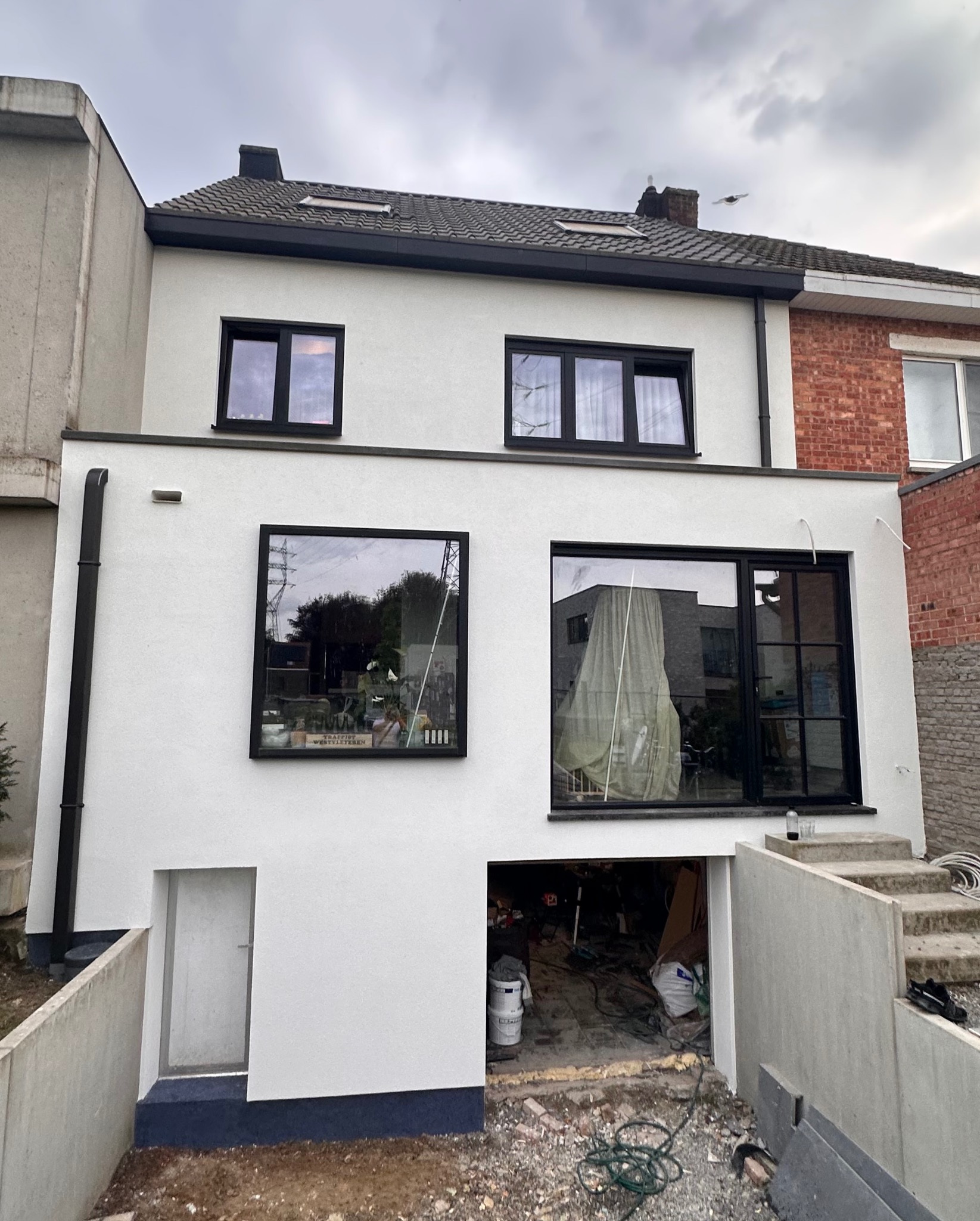 Dwupiętrowy dom z nową, białą elewacją i czarnymi ramami okiennymi, widok na częściowo wykończony teren wokół domu, z otwartym garażem ukazującym narzędzia i materiały budowlane.