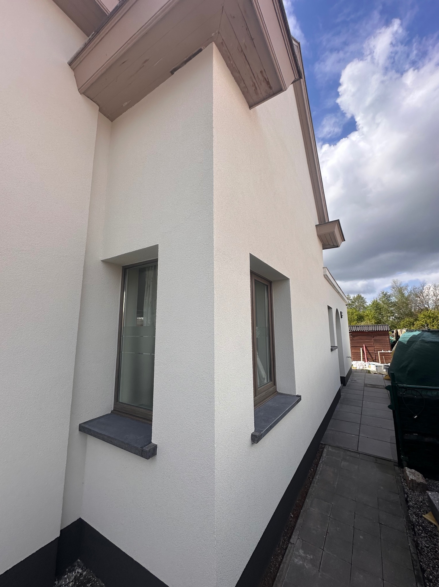 Boczny widok domu z nową, jasną elewacją o grubej fakturze, z szarymi parapetami i ciemną podmurówką, na tle błękitnego nieba z chmurami.