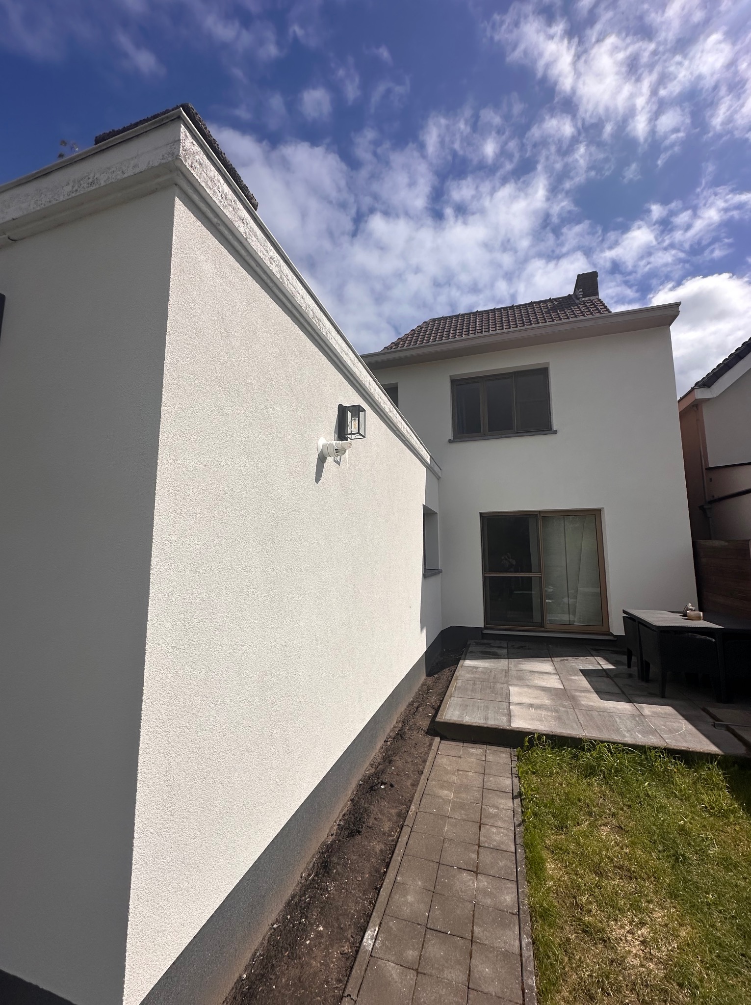 Odnowiona elewacja domu jednorodzinnego w jasnym odcieniu, widok z boku na ścianę z oświetleniem zewnętrznym i fragment tarasu z meblami ogrodowymi.