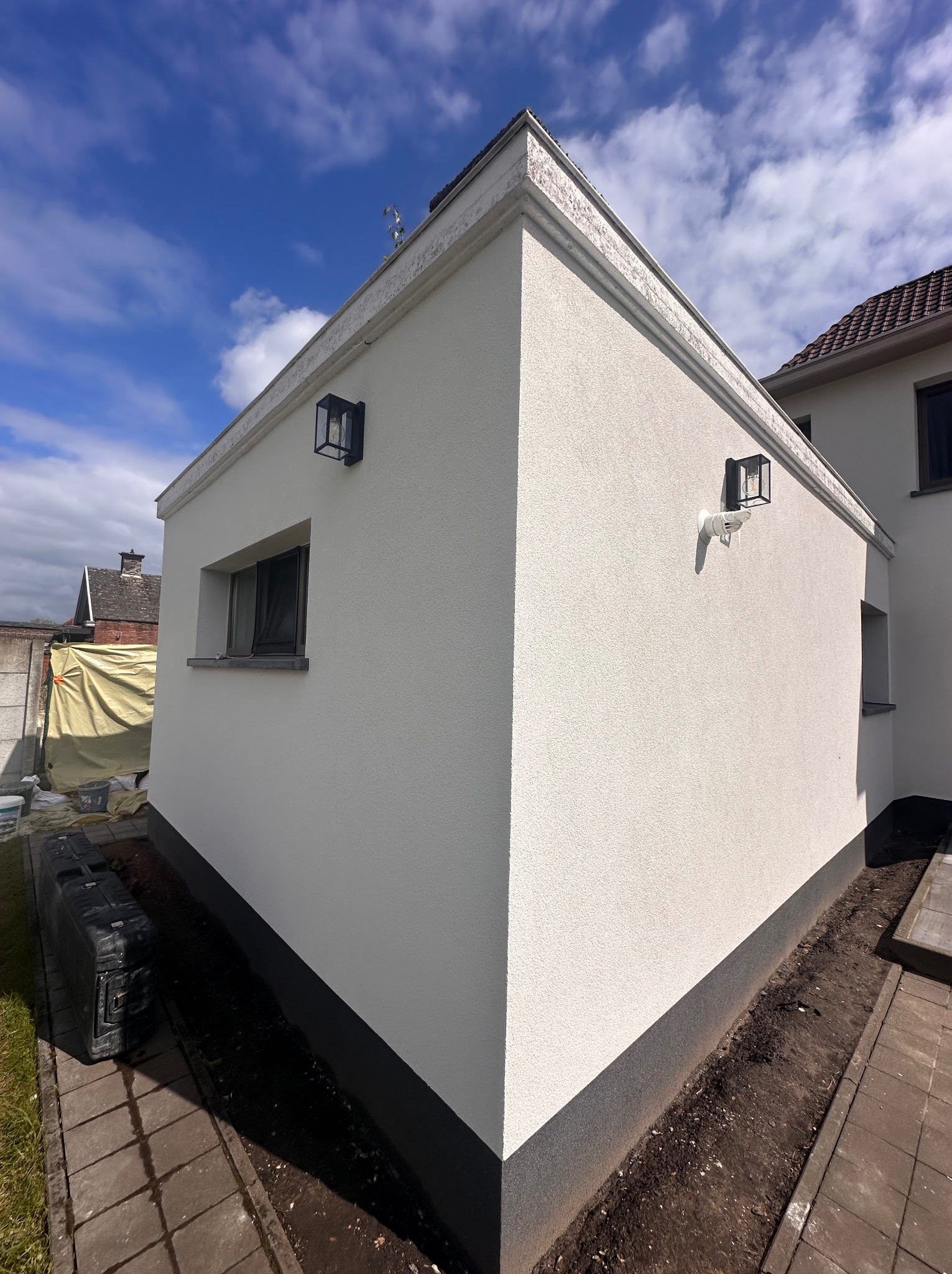 Wykończona elewacja budynku z białym tynkiem strukturalnym, czarną podmurówką i lampami ściennymi w stylu industrialnym, widok z boku na tle błękitnego nieba z chmurami.