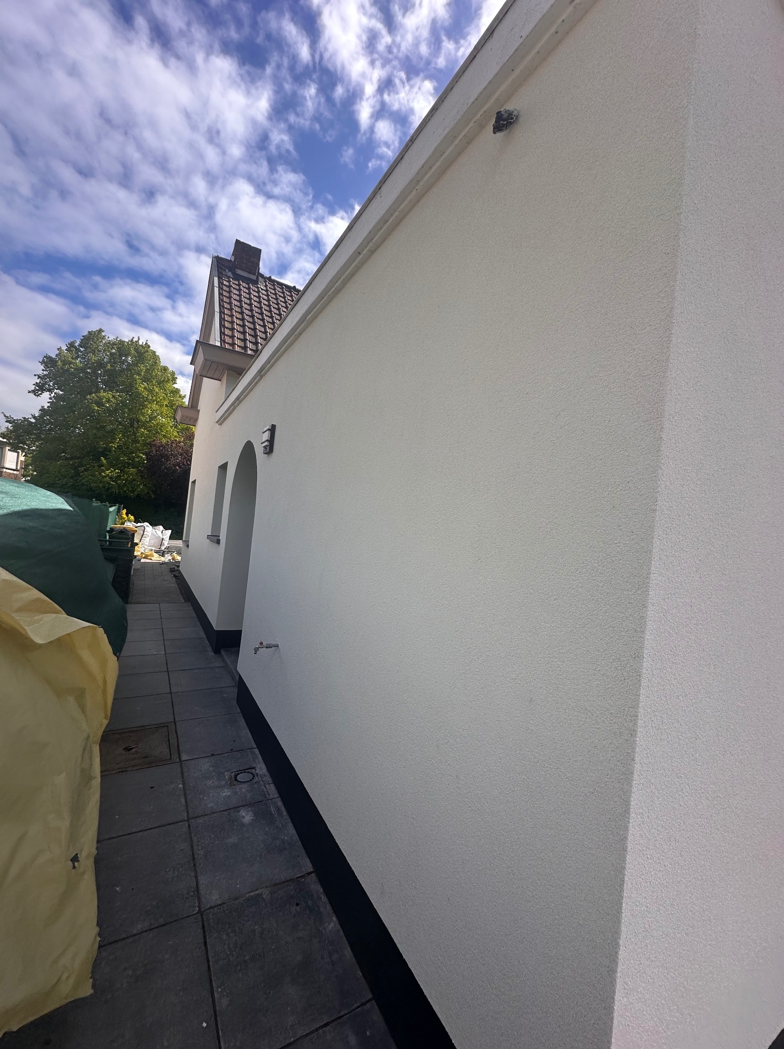 Świeżo otynkowana elewacja budynku z widoczną fakturą, z fragmentem dachu pokrytego czerwoną dachówką, błękitnym niebem z chmurami w tle oraz fragmentem zadaszenia z workami budowlanymi po lewej...