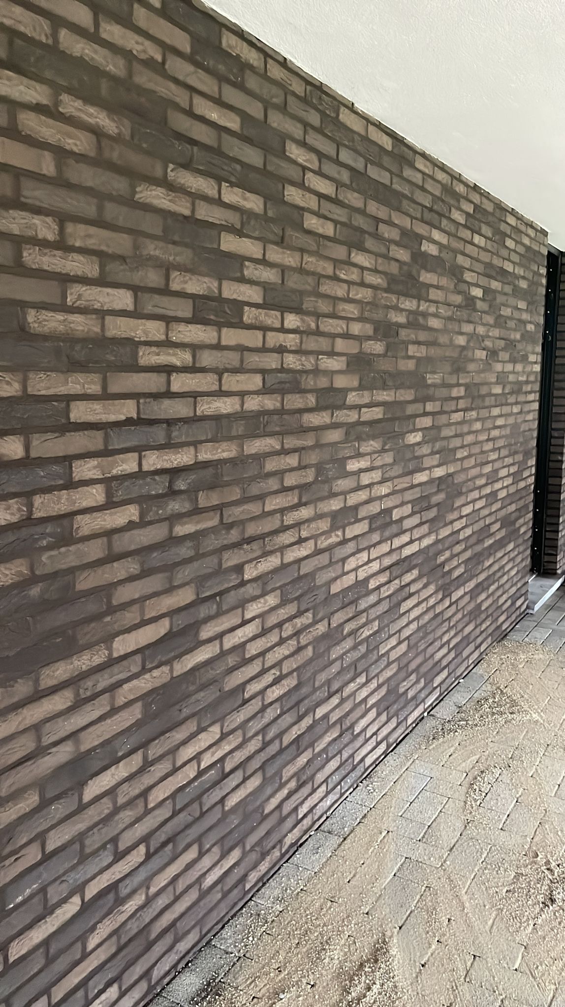 Fragment ściany zewnętrznej budynku wykończonej brązową cegłą klinkierową z widocznymi fugami w odcieniach szarości, z drobnymi pozostałościami piasku na chodniku.