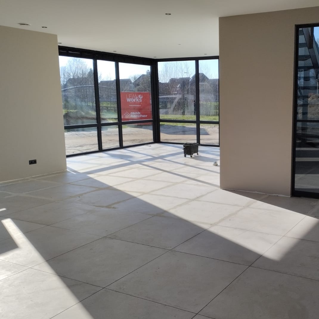 Jasne wnętrze z ułożoną, kwadratową podłogą z płytek w odcieniach szarości, duże okna z czarnymi ramami wpuszczają dużo światła, widoczna reklama na zewnątrz, w rogu stoi mały wózek narzędziowy.